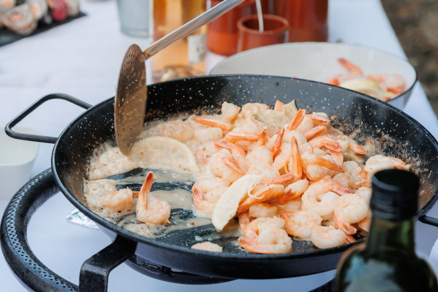 Fresh shrimp being cooked in a large black paella pan on an outdoor gas stove at a catered event.