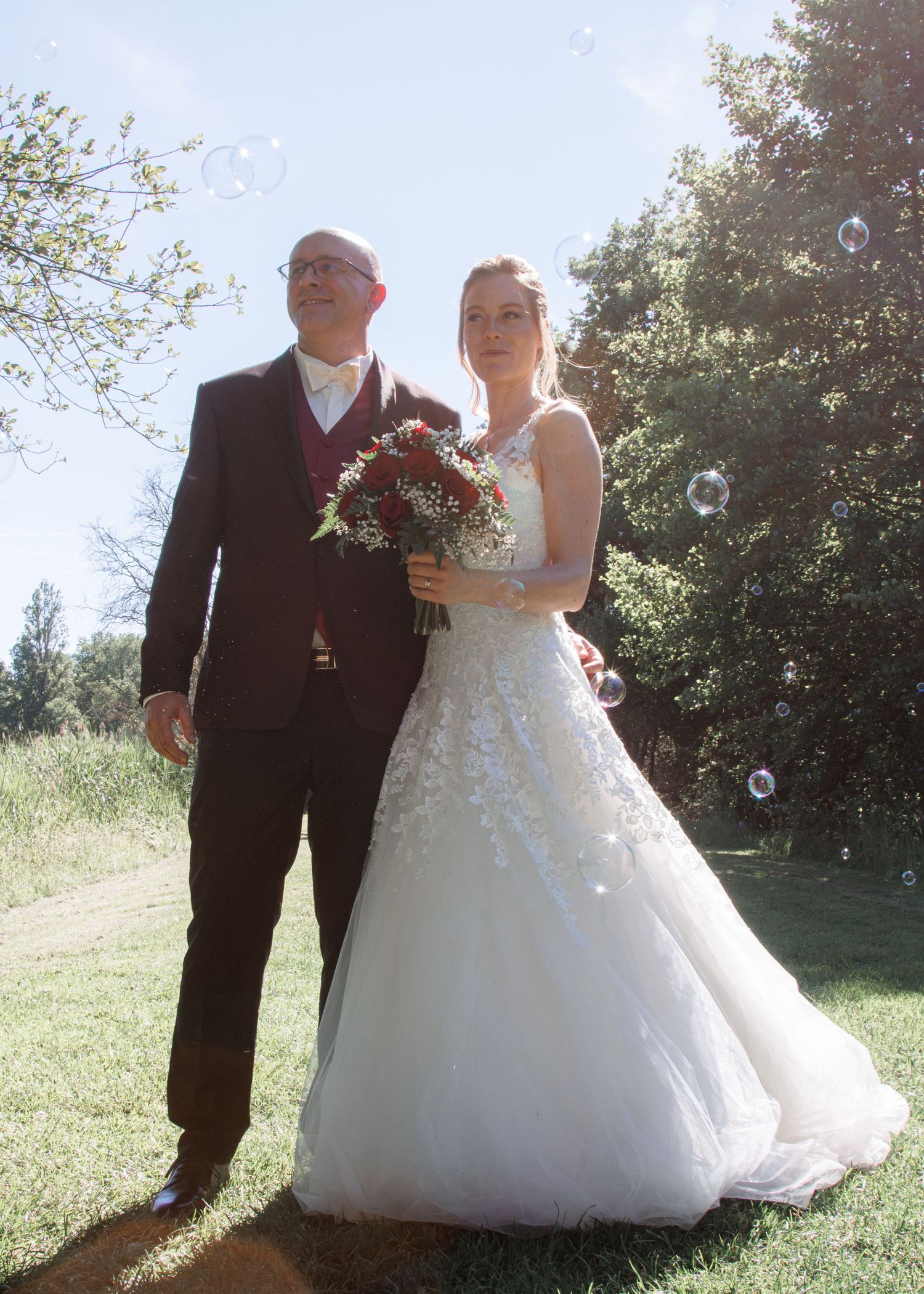 Wedding couple poses outdoors in sunlight with soap bubbles floating around them, bride holding red bouquet.