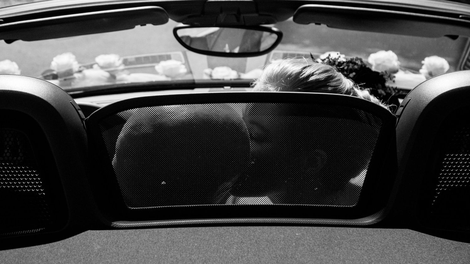 Black and white photo of newlyweds sharing a kiss viewed through a car rear window.