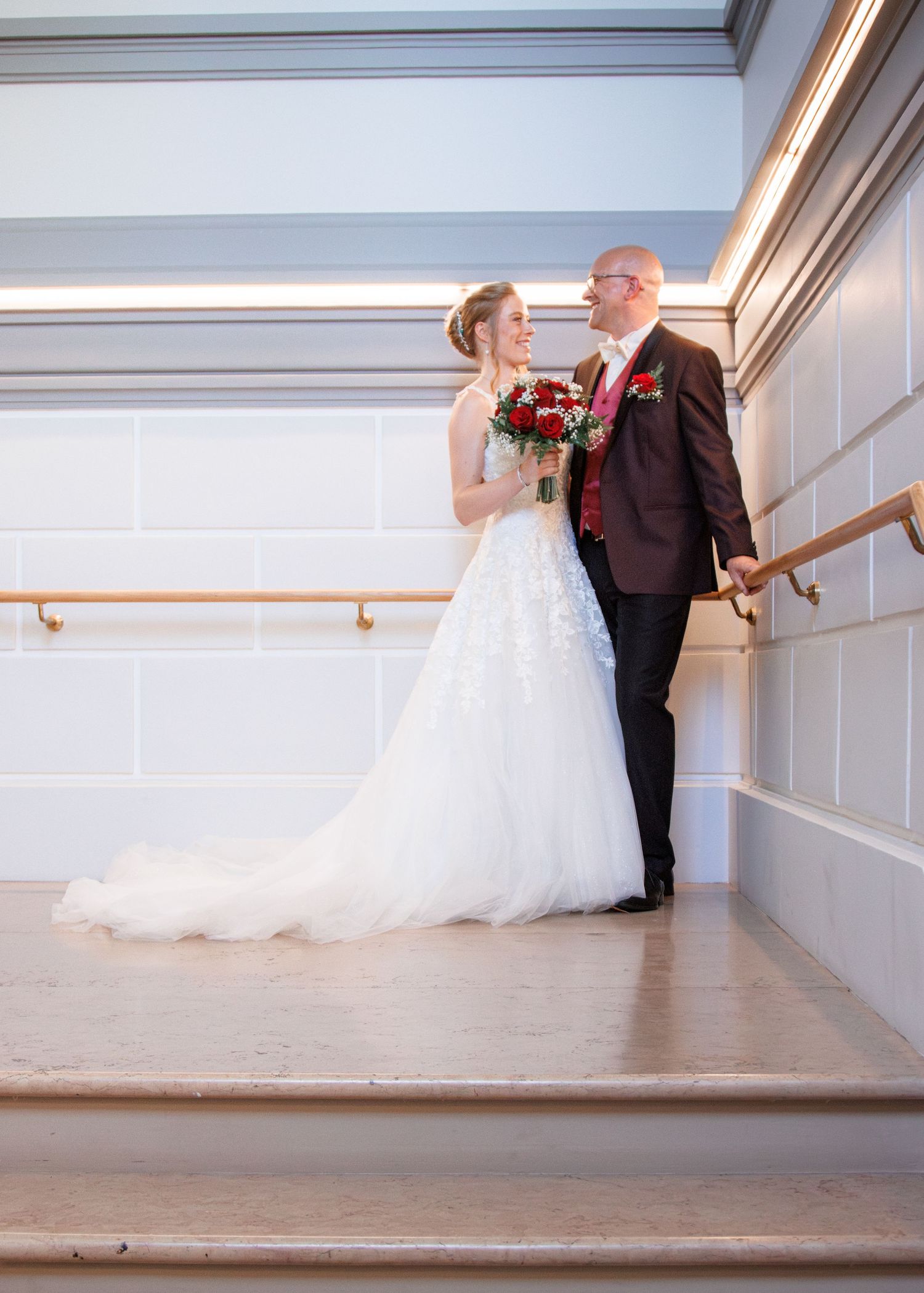 Bride and groom share a romantic moment on elegant staircase, bride holding red bouquet in white gown.