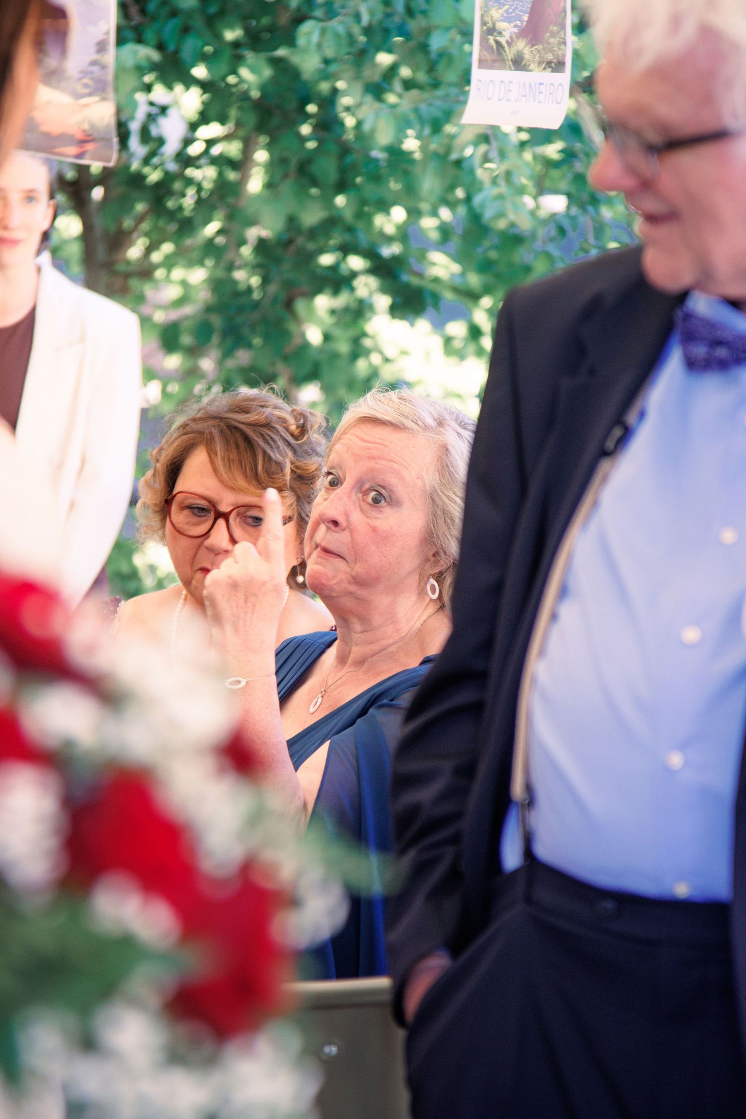 Two older women at wedding reception, one wiping tears, surrounded by guests and red flowers.