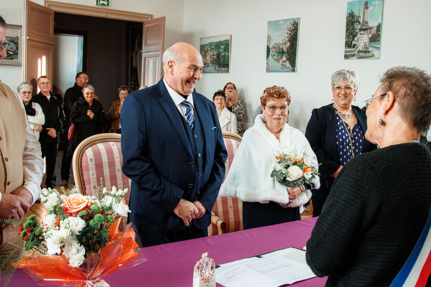 Elderly couple smiling during an intimate civil wedding ceremony indoors, with guests and floral arrangements in the background.