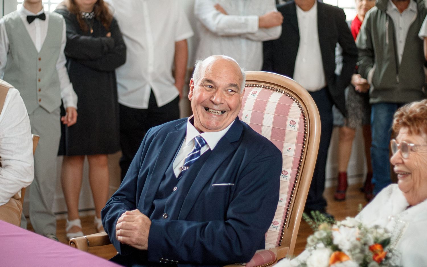 Smiling bald man in navy suit sitting in ornate chair at a wedding ceremony with guests standing behind.