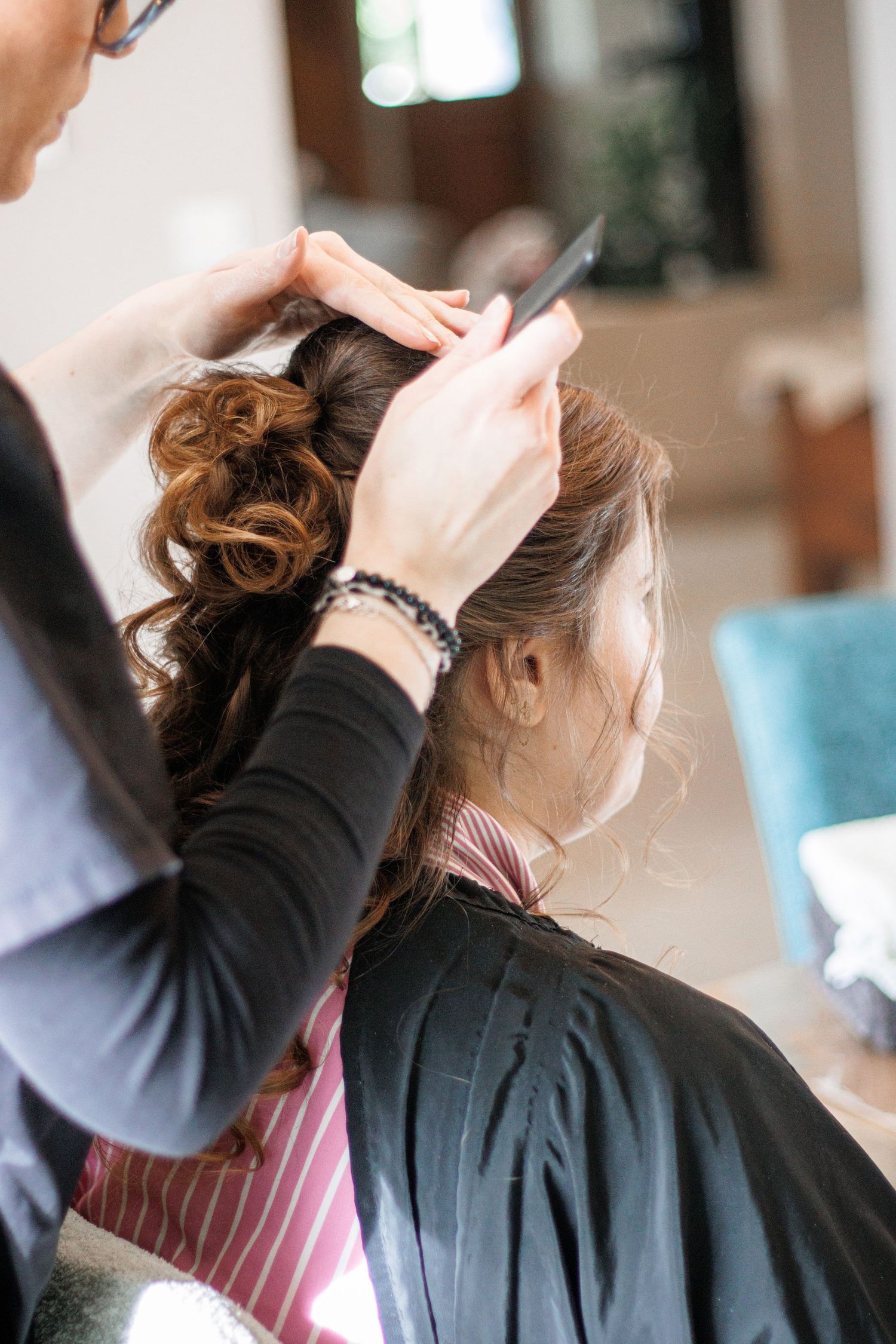 A hairstylist carefully styling a woman's curly updo with a comb in a bright salon setting.