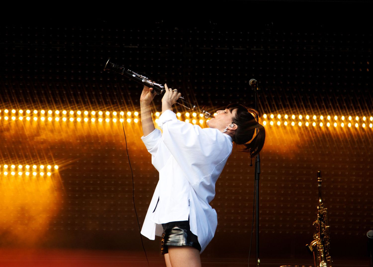 Performer in white shirt plays clarinet on stage with warm golden lights backdrop at live concert.
