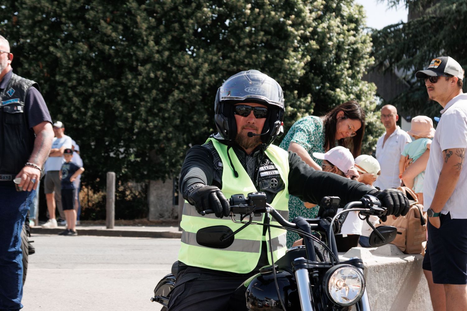 Motorcyclist in helmet and safety vest rides at outdoor event with spectators watching in background.