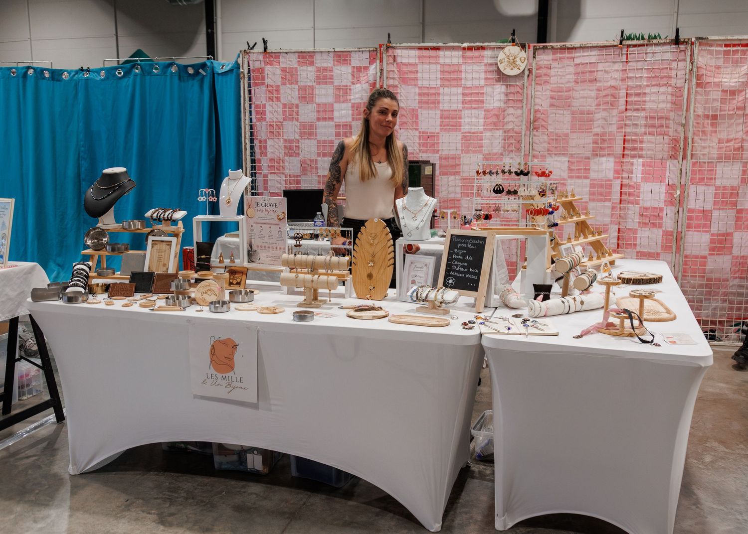 Woman standing behind a craft fair booth displaying handmade jewelry and wooden accessories on white draped tables.