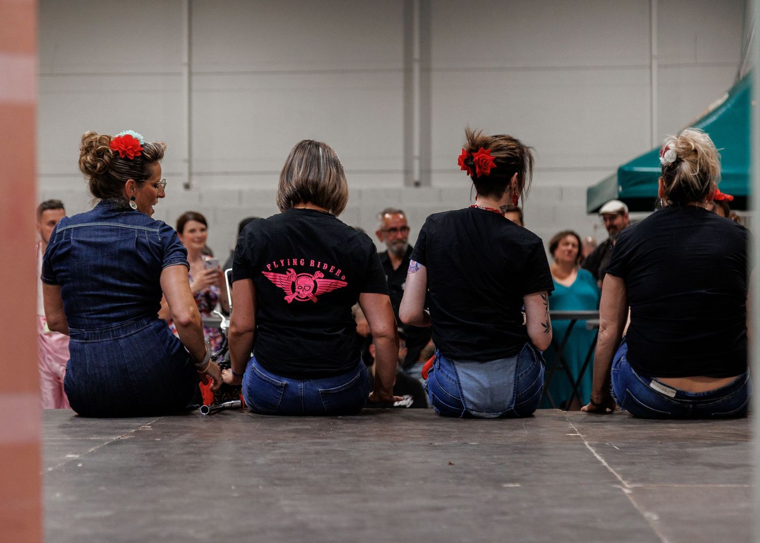 Women in matching black shirts with red floral hair accessories sit together on a floor at an indoor event.