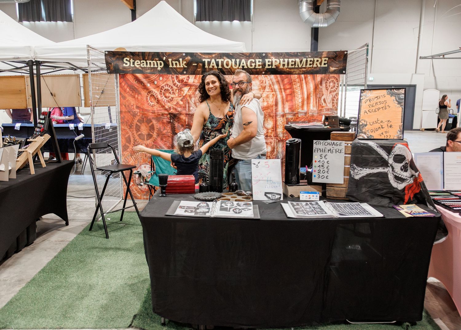 Steamp Ink temporary tattoo booth with vendors posing at a black draped table at an indoor market event.