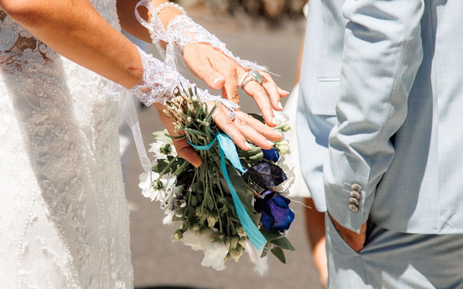 Close-up of hands holding a wedding bouquet with blue and white flowers tied with teal ribbon.