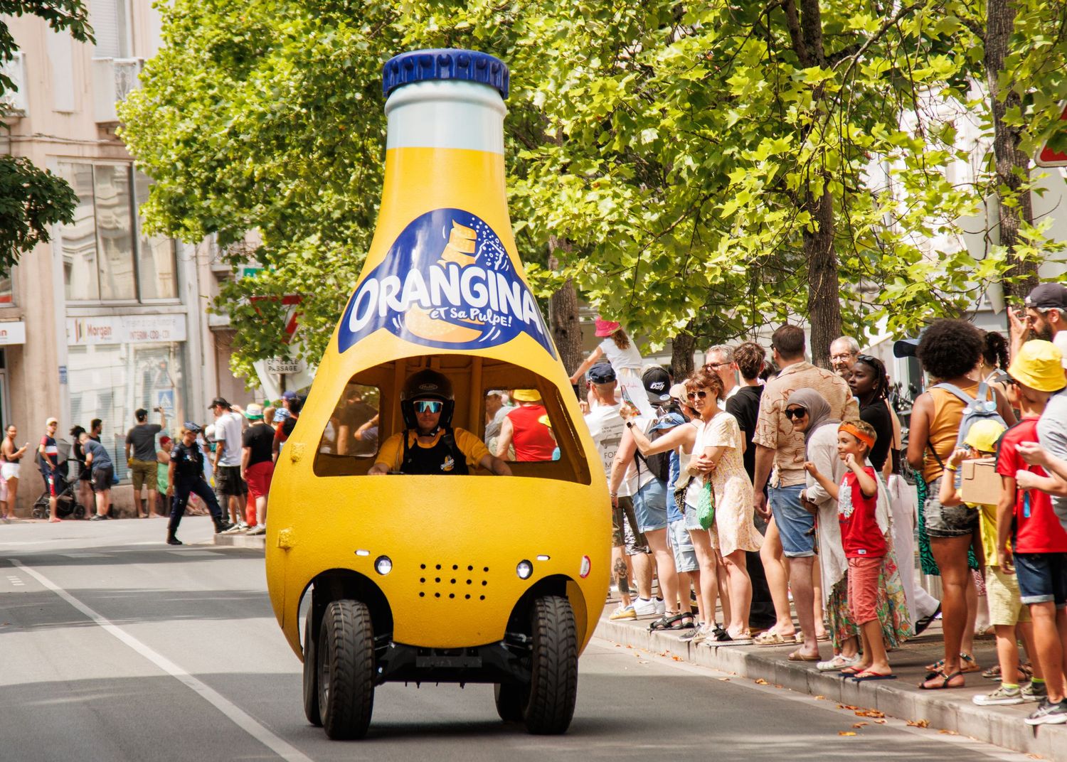 A unique yellow promotional vehicle shaped like a bottle driving through a crowded street.