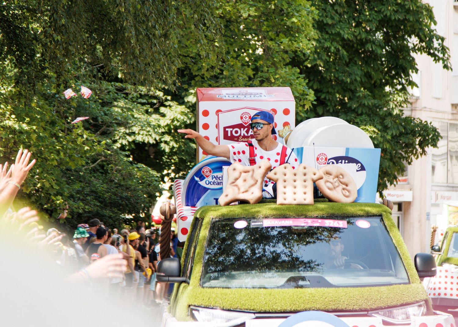 Promotional vehicle decorated with Tour de France merchandise driving through tree-lined street.