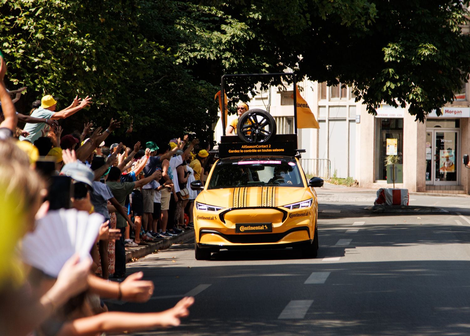 A yellow promotional vehicle parades through streets as crowds wave from both sides.