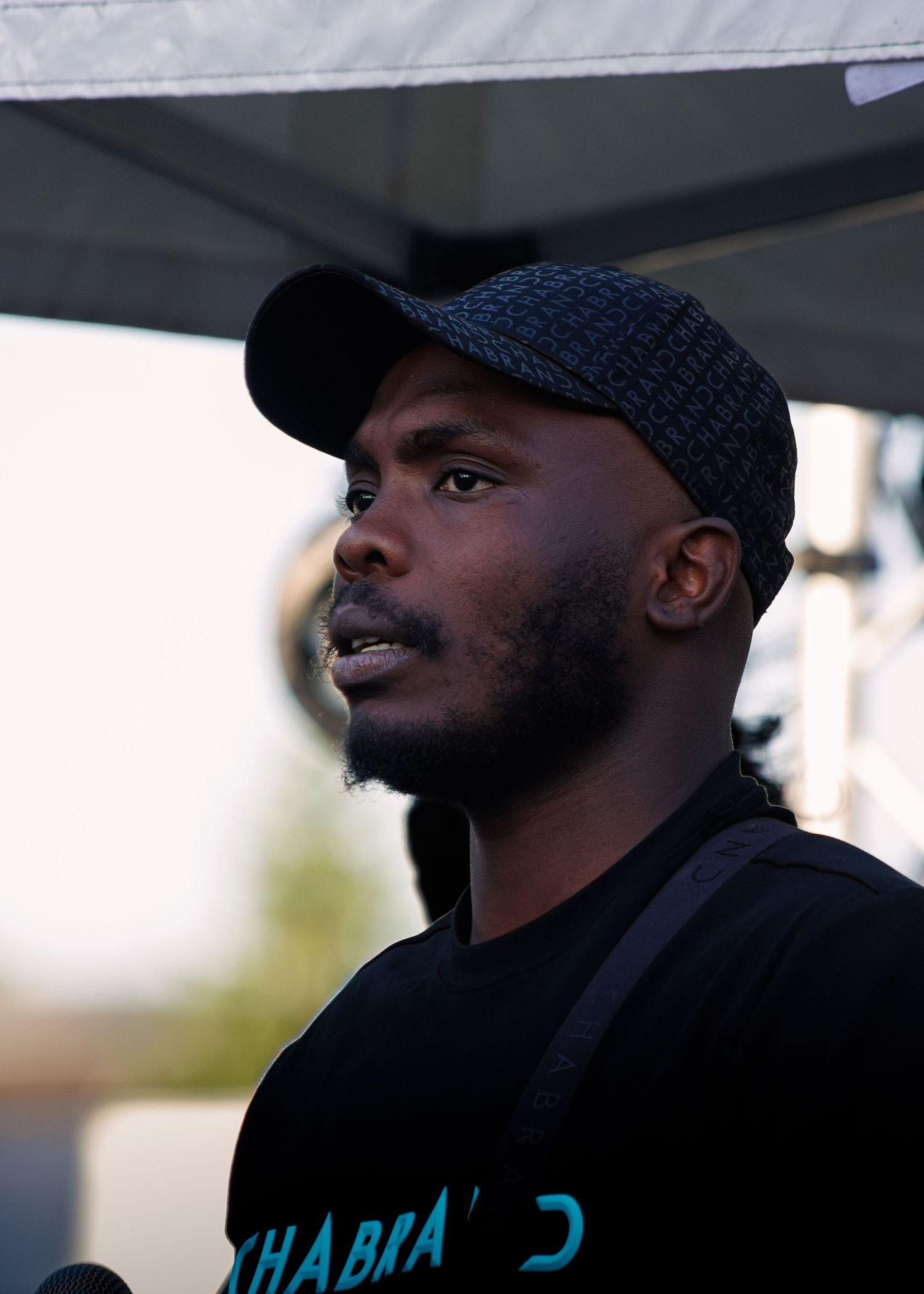 Person in black hat and clothing speaks into microphone.