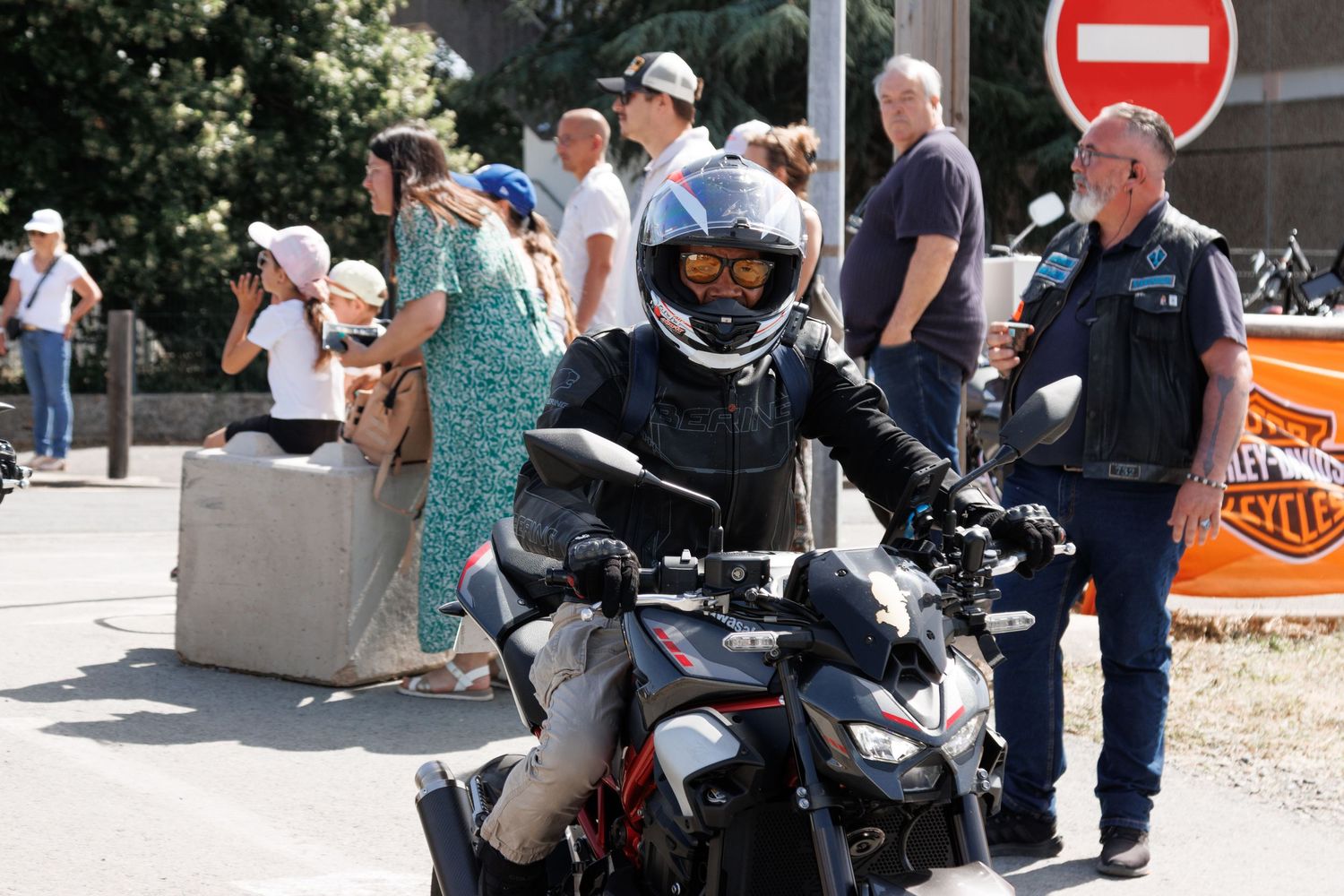 A motorcyclist in a helmet rides past spectators near a no-entry sign at an outdoor Harley-Davidson event.