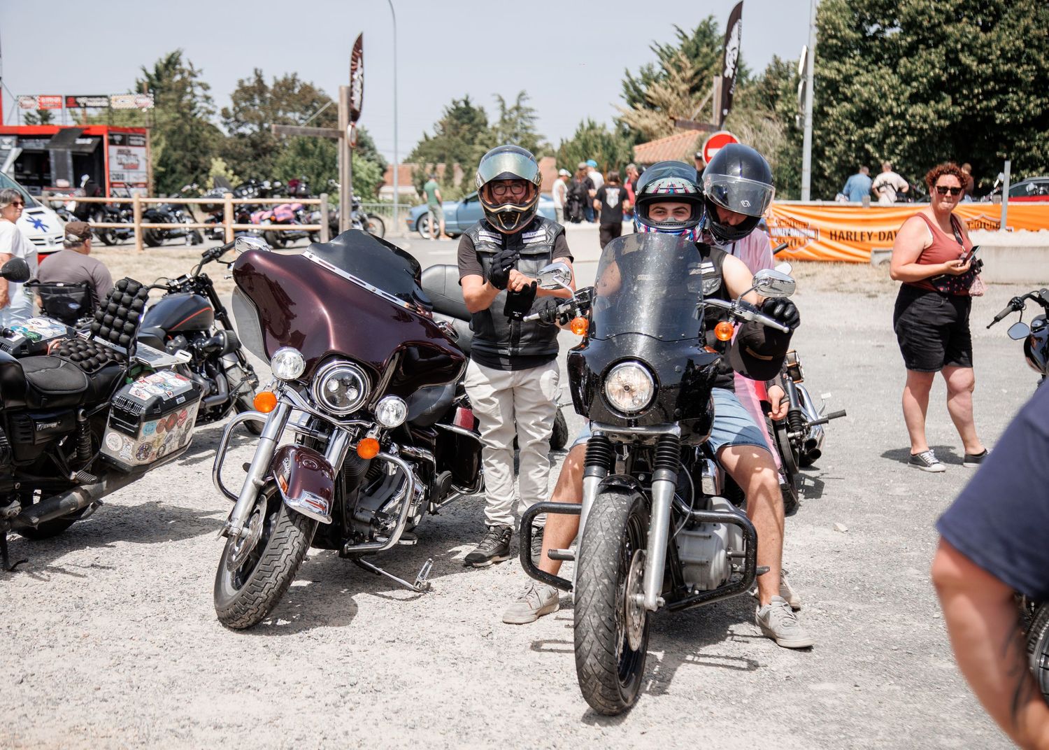 Motorcyclists riding Harley-Davidson bikes at an outdoor rally on a sunny day, with spectators watching nearby.