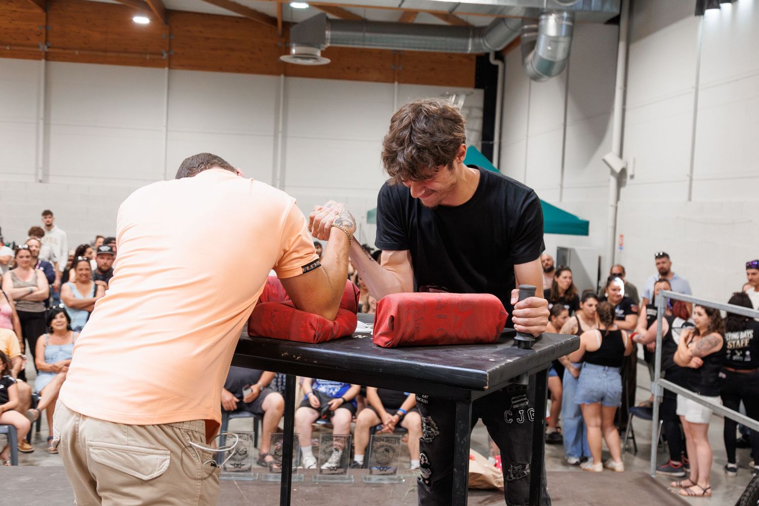 Two men competing in an arm wrestling match at a public event with a crowd of spectators watching in the background.