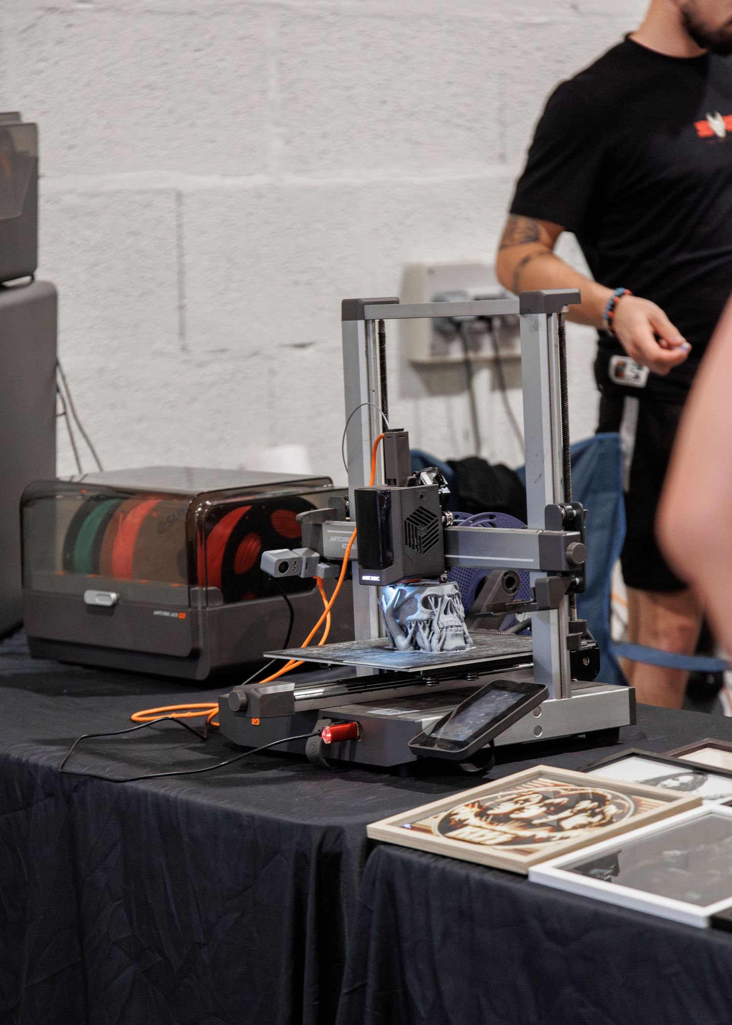 A 3D printer on a black table at a maker fair, with printed objects nearby and a tattooed person standing behind.