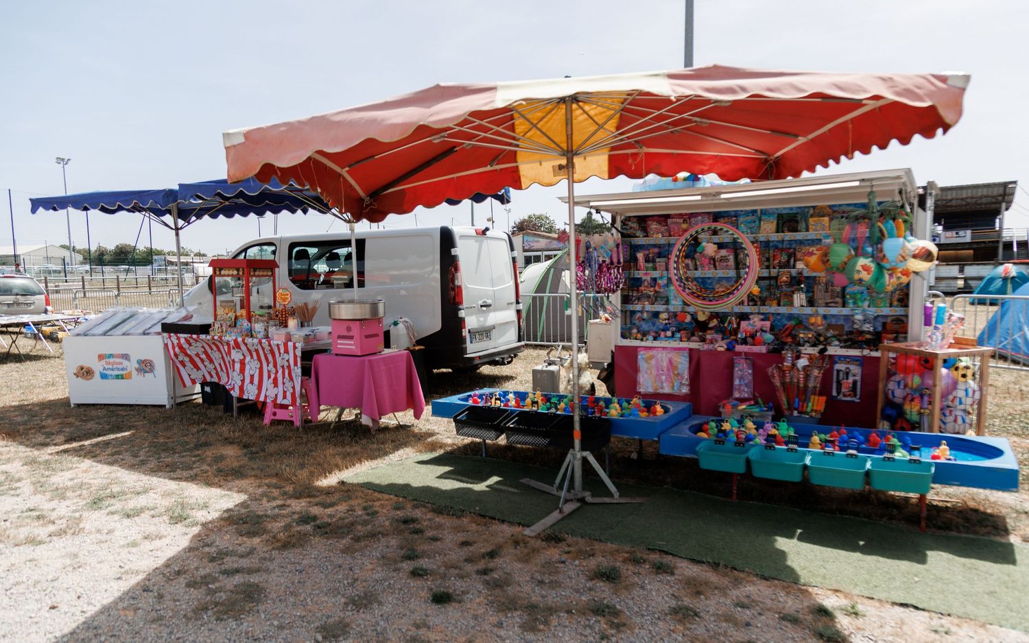 Colorful carnival game booths with red striped awnings set up outdoors on sandy ground at a fairground.
