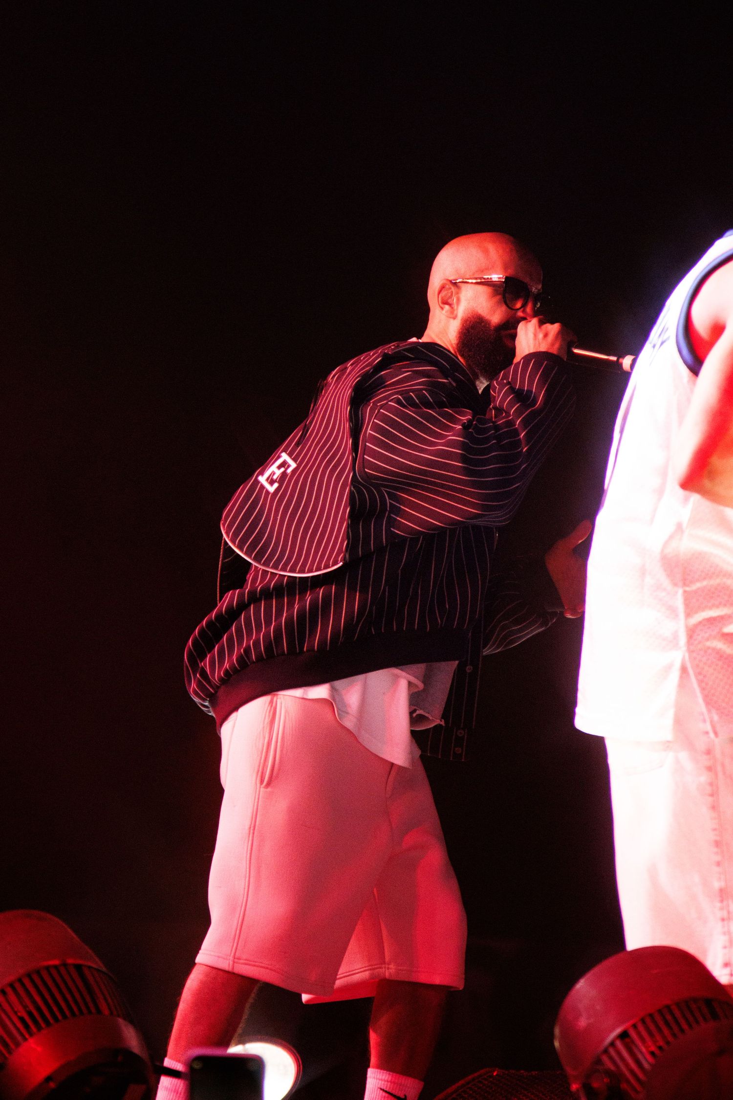 A performer in a striped shirt and white shorts sings into a microphone on stage under dramatic red lighting.