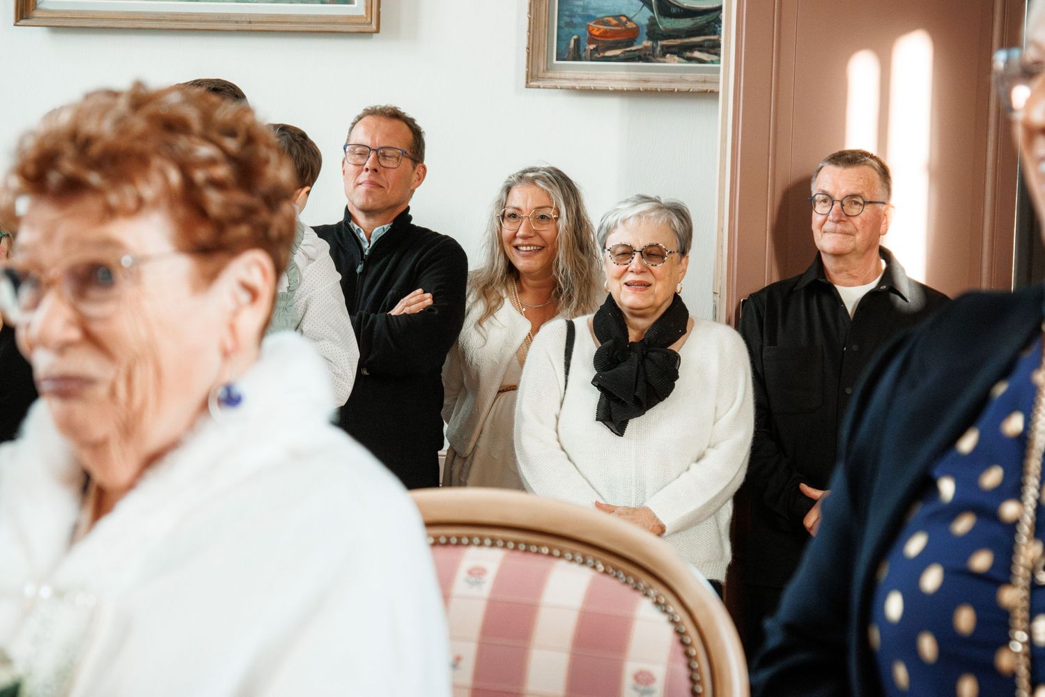 Group of smiling adults gathered indoors at a social event, with artwork visible on the wall behind them.