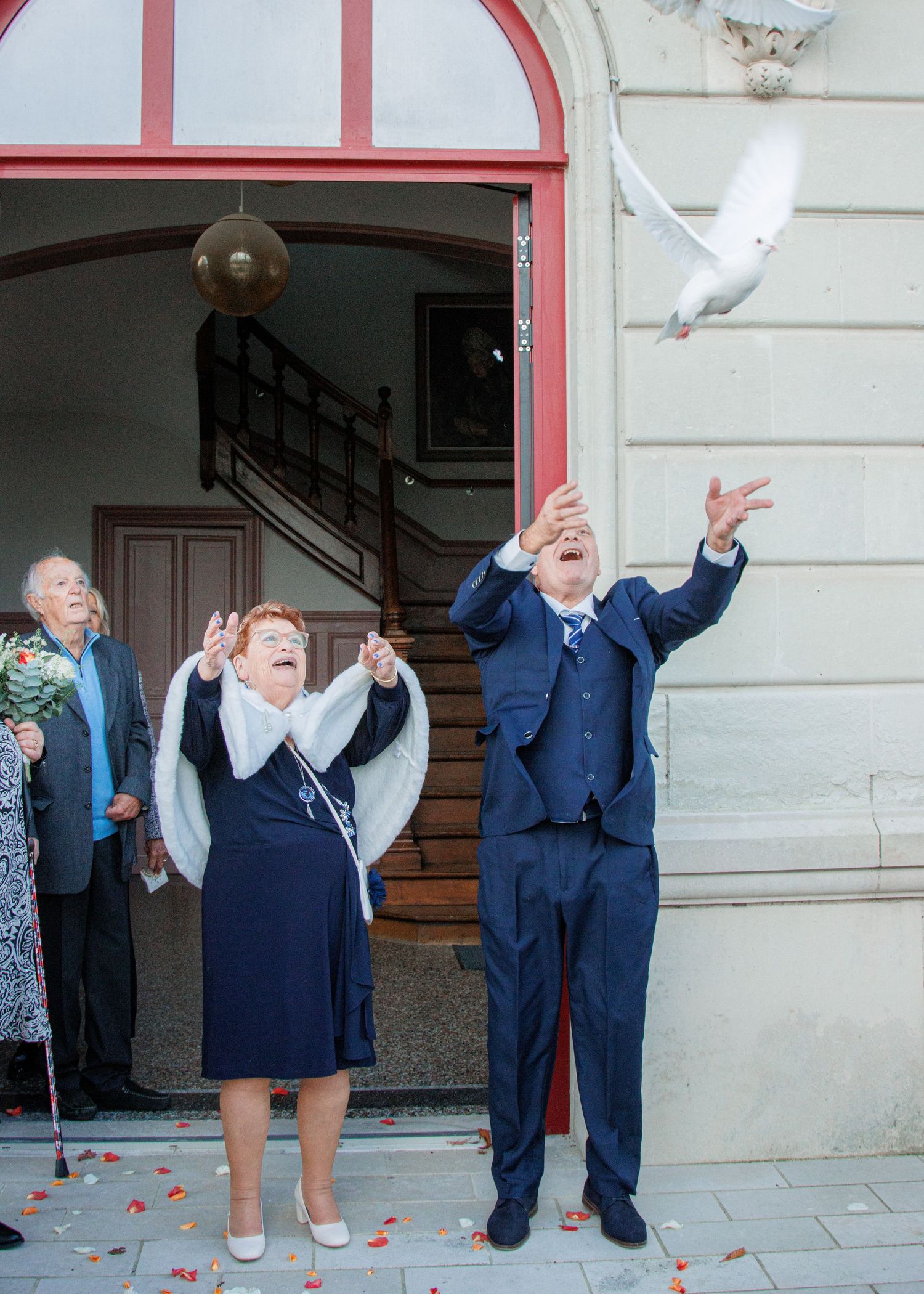 Elderly couple releasing white doves outside a building entrance during a wedding ceremony celebration.