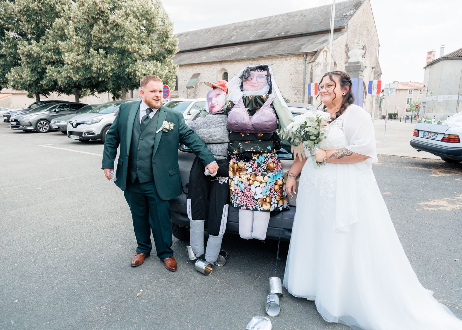 Laughing bride and groom posing with a funny stuffed dummy decoration attached to their wedding car.
