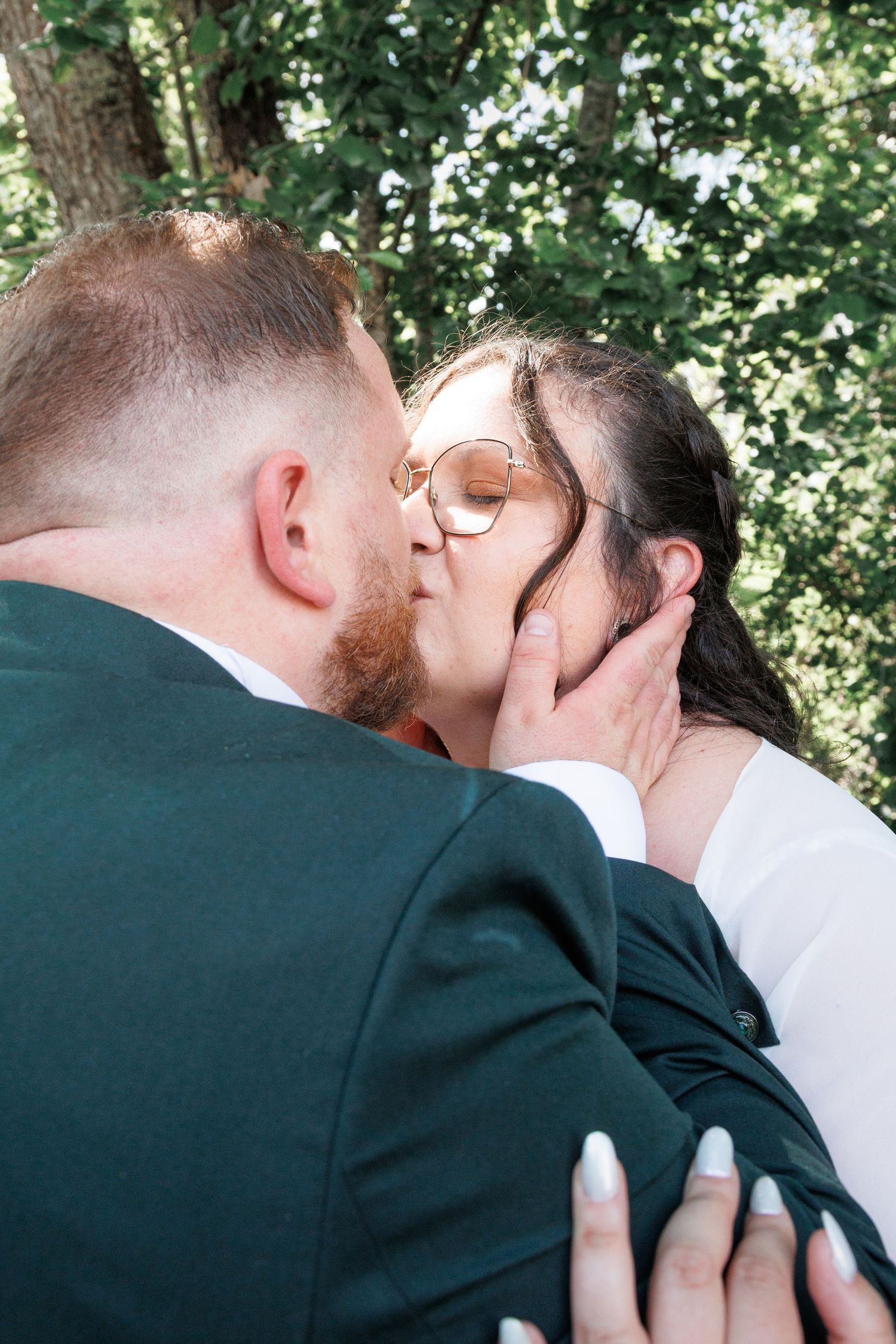 Newlyweds share a close romantic kiss outdoors surrounded by lush green foliage.