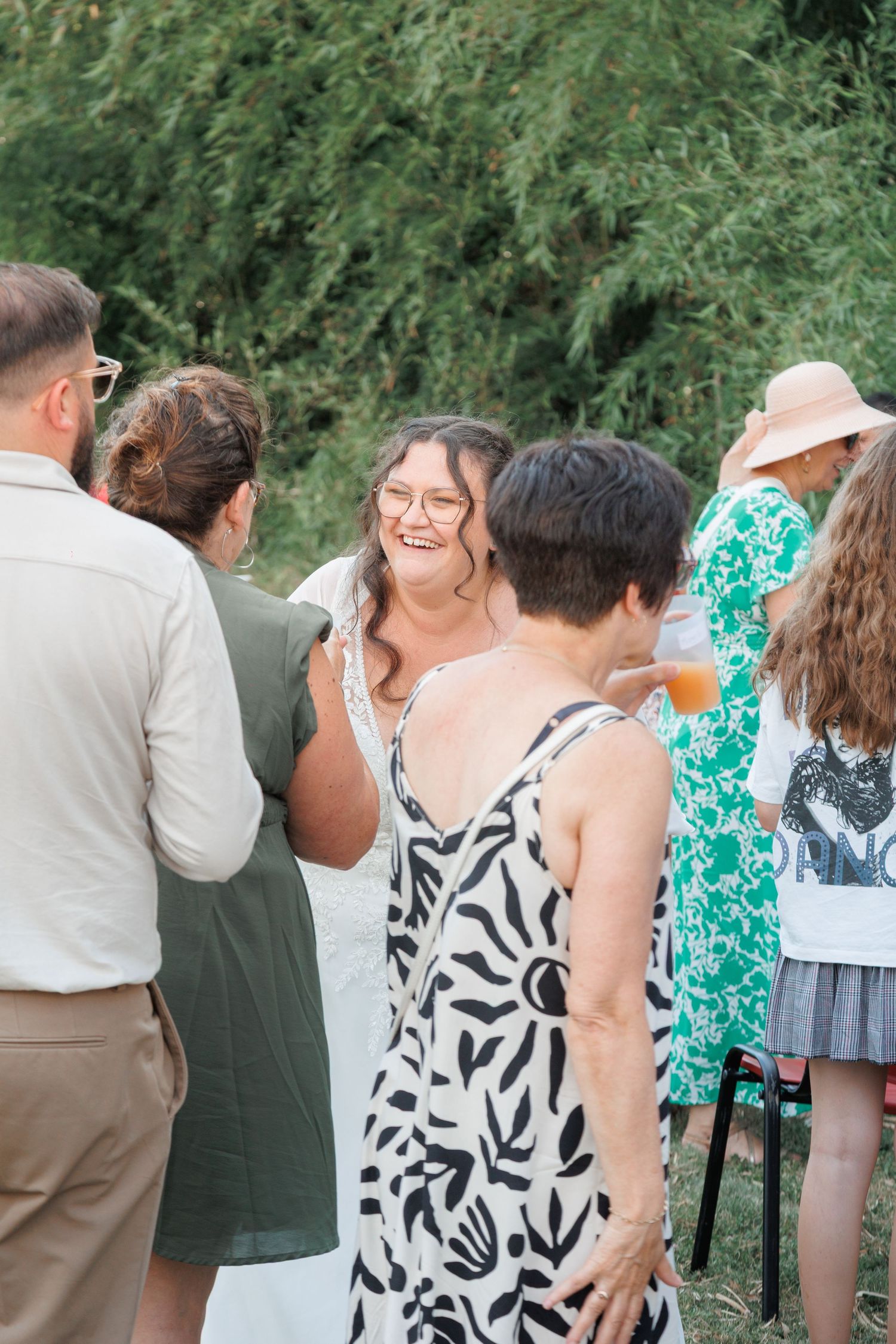 Smiling woman in white lace dress laughs while socializing with guests at an outdoor wedding cocktail hour.