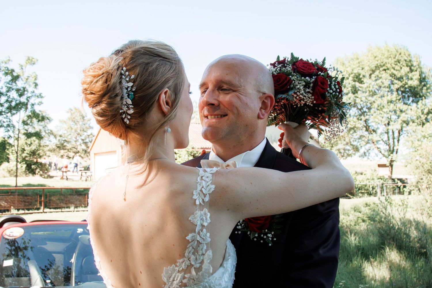 Smiling bald groom embraced by bride holding red bouquet on their wedding day outdoors.