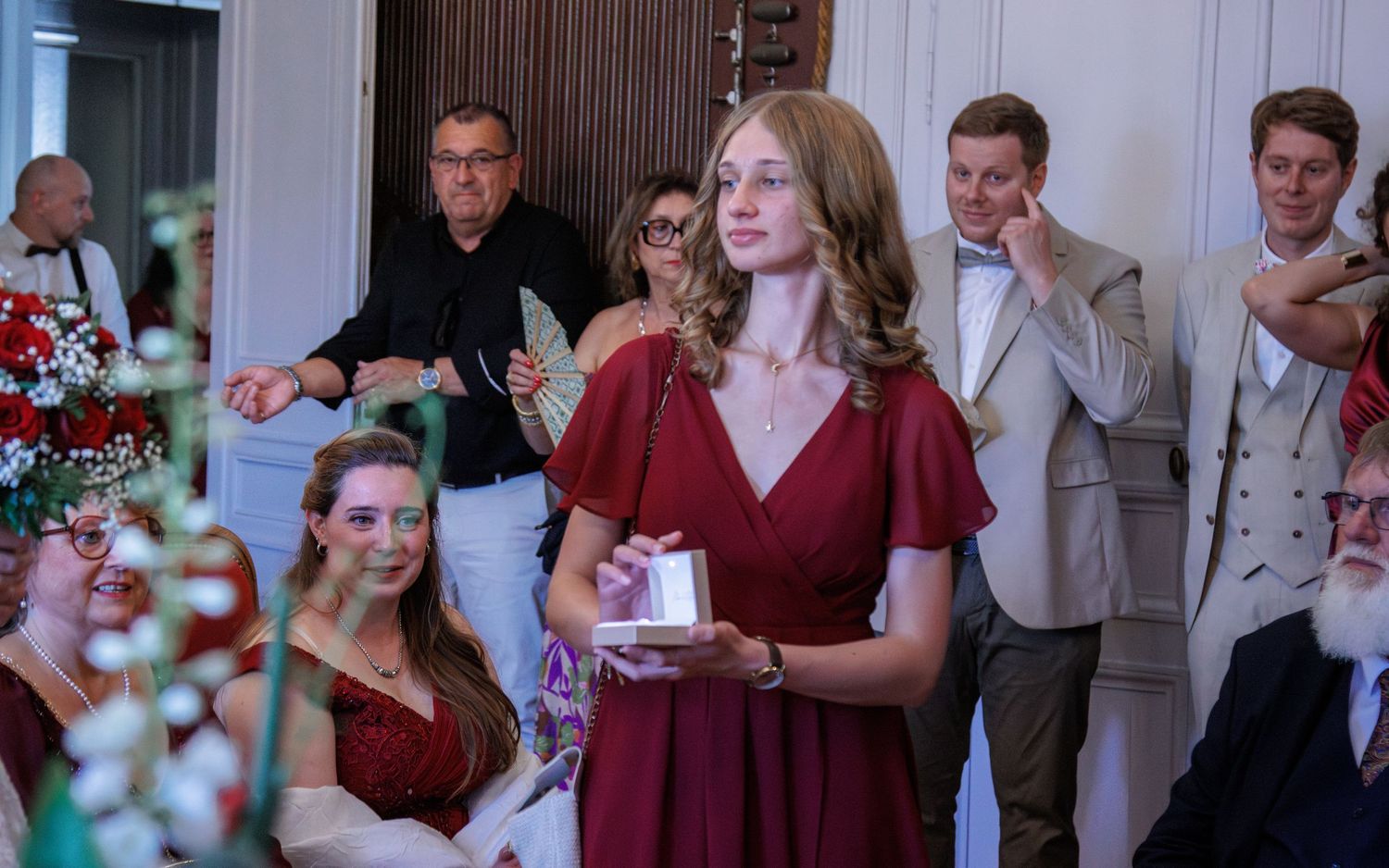 Young woman in red dress holds gift box during indoor wedding celebration.