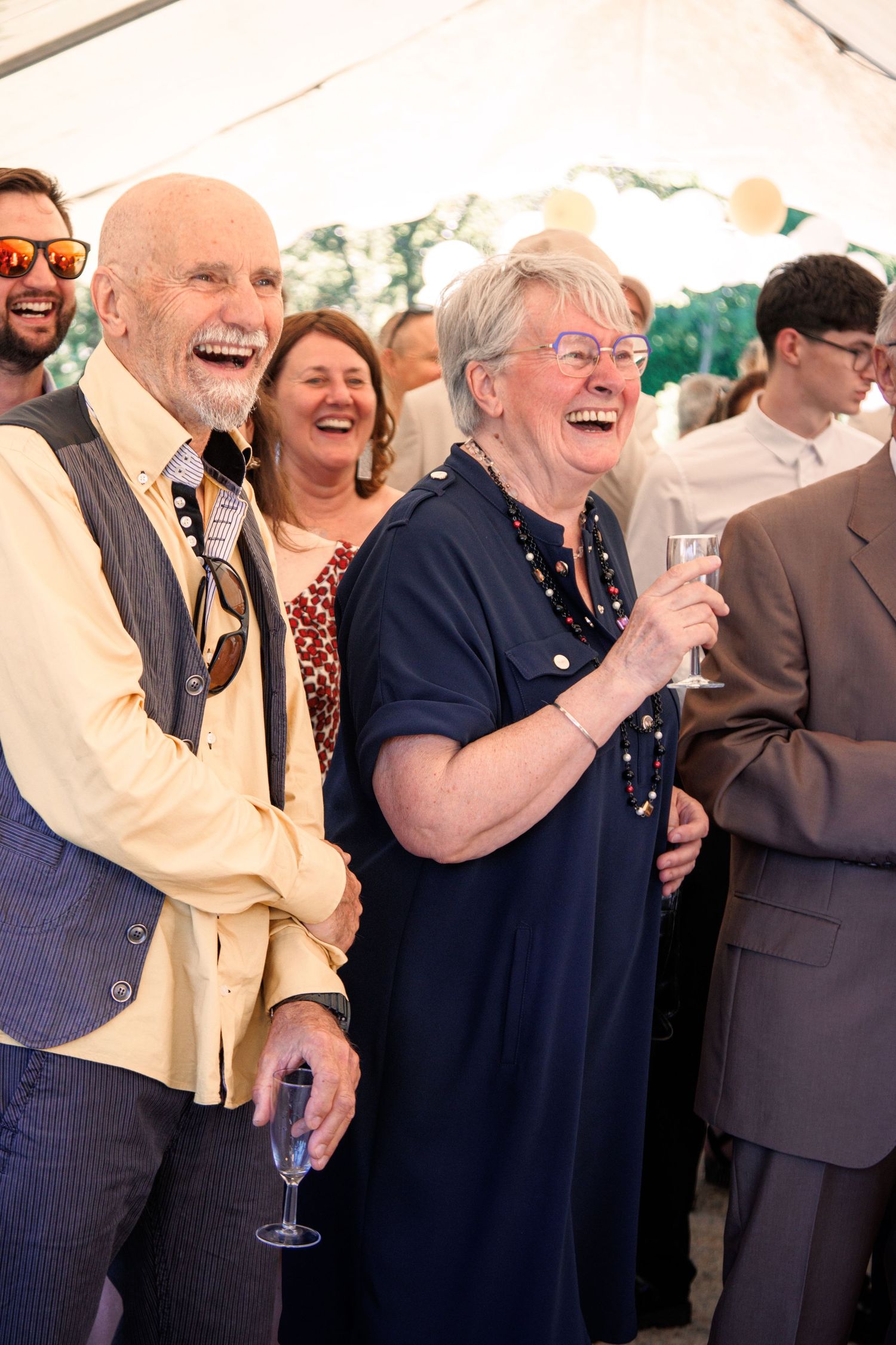 Joyful elderly wedding guests laughing and holding champagne glasses under a marquee tent.