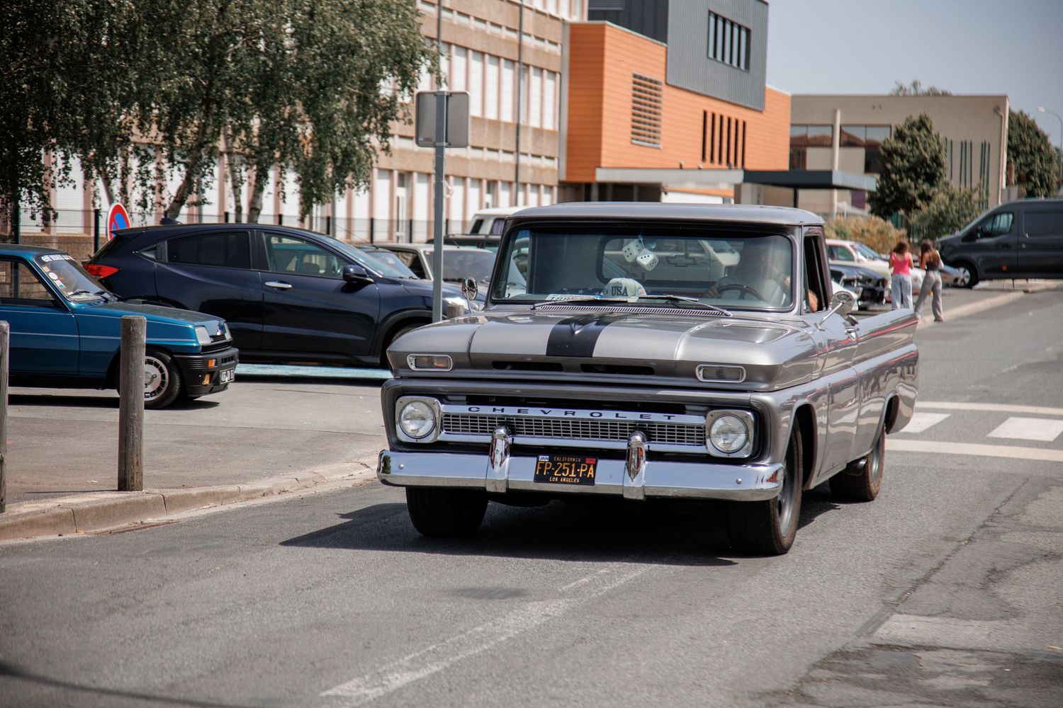 Vintage silver Chevrolet pickup truck driving on a sunny street with parked cars and an orange building in the background.