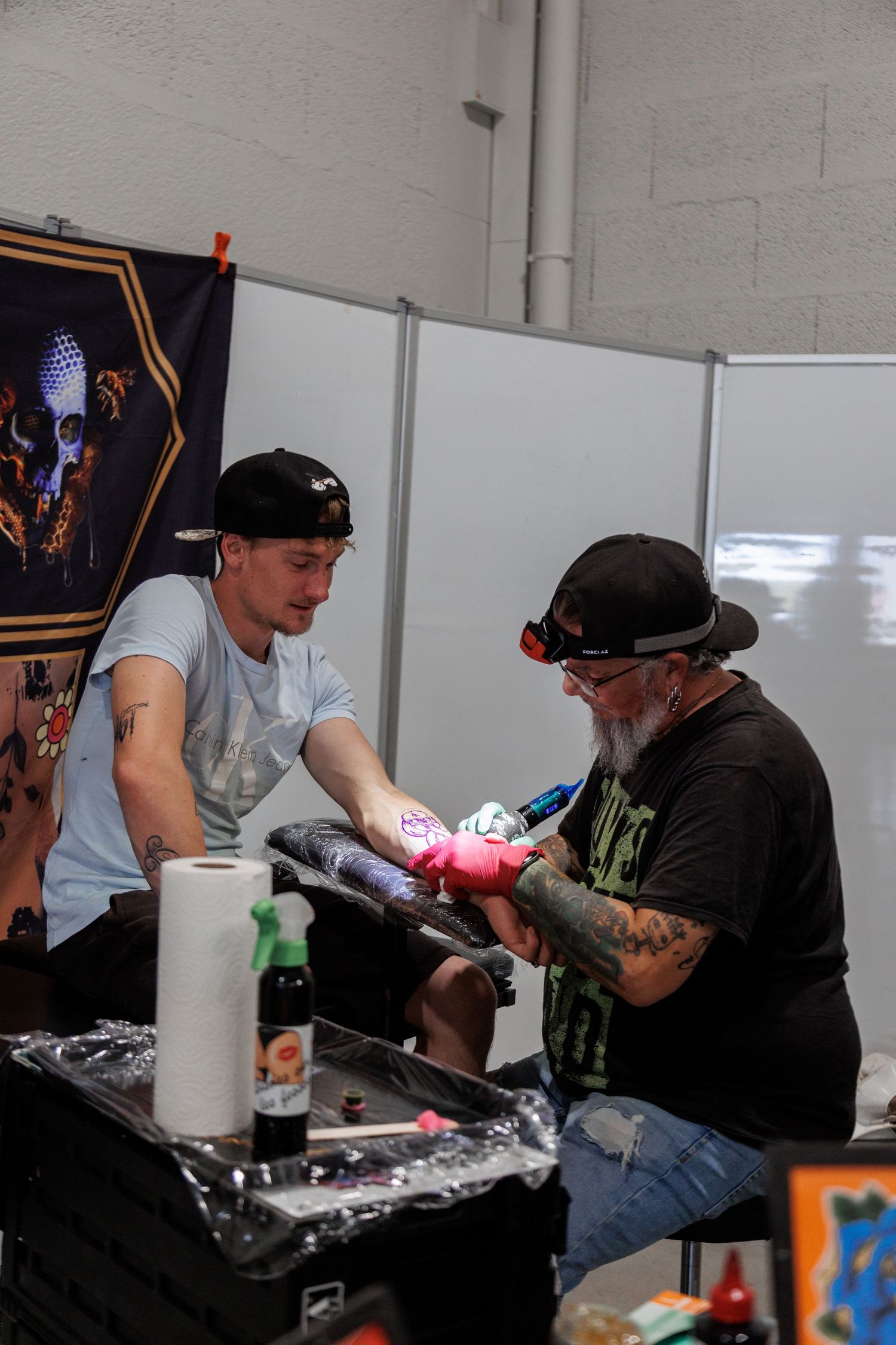 Tattoo artist with beard and cap tattooing a client's arm at a convention booth with skull artwork in background.