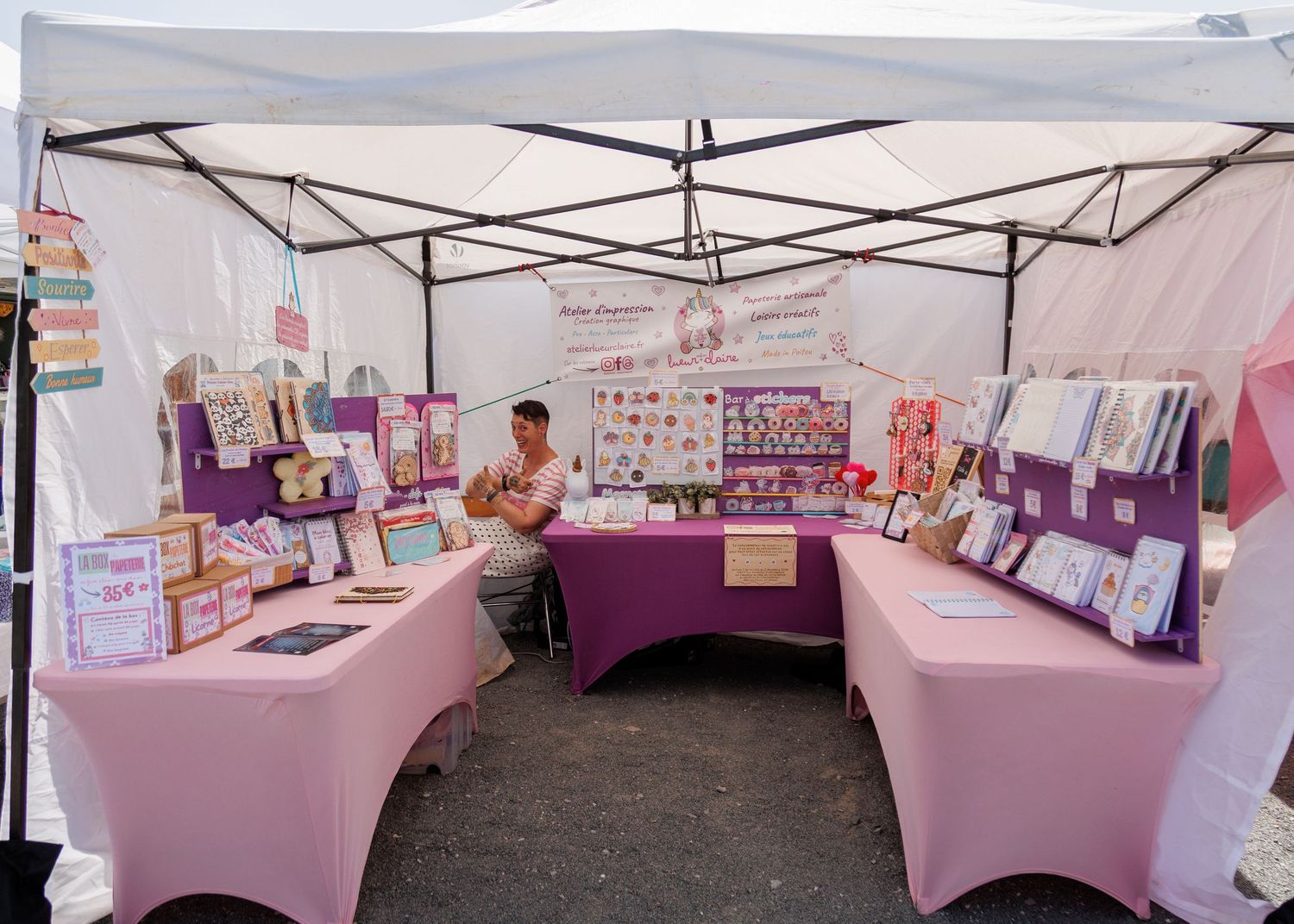Outdoor craft fair booth with pink tables displaying handmade jewelry and accessories under a white canopy tent.