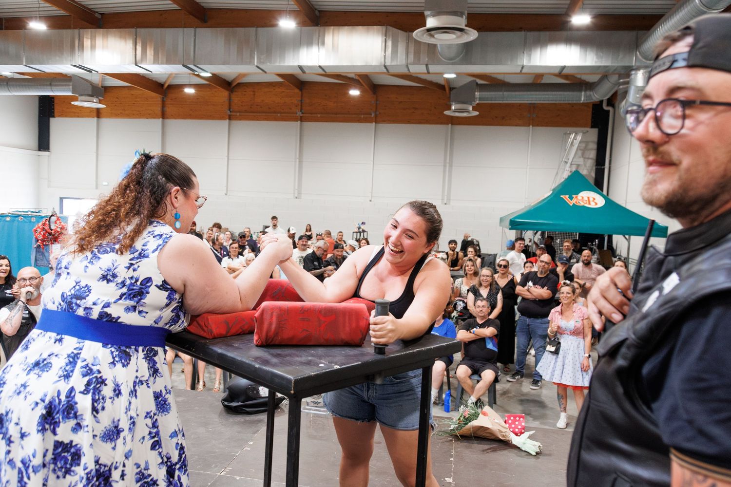 Two women arm wrestling on stage at an indoor event with a crowd of spectators watching in the background.