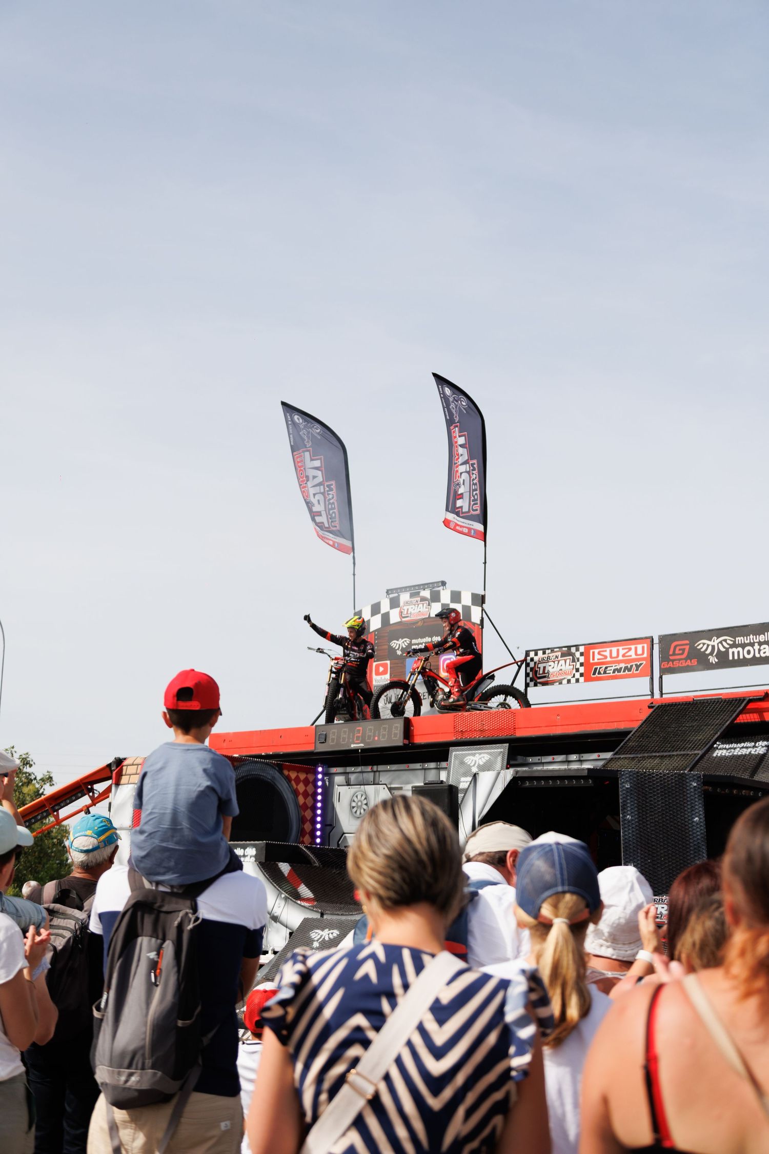 Motorcycle stunt riders perform on elevated stage at outdoor motorsport event with crowd watching and branded flags waving.