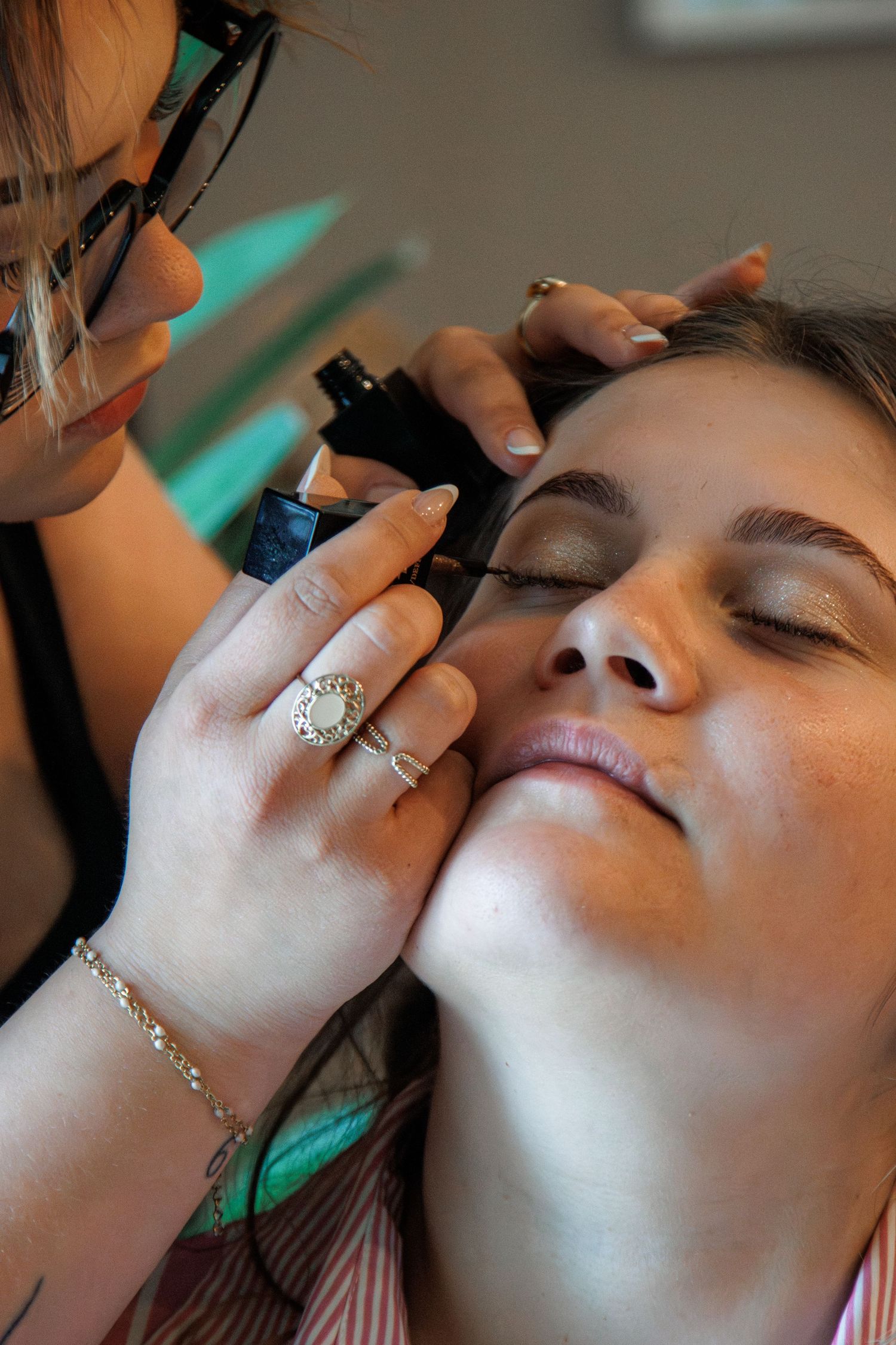 A makeup artist applying mascara to a woman with closed eyes, wearing a diamond ring and bracelet.