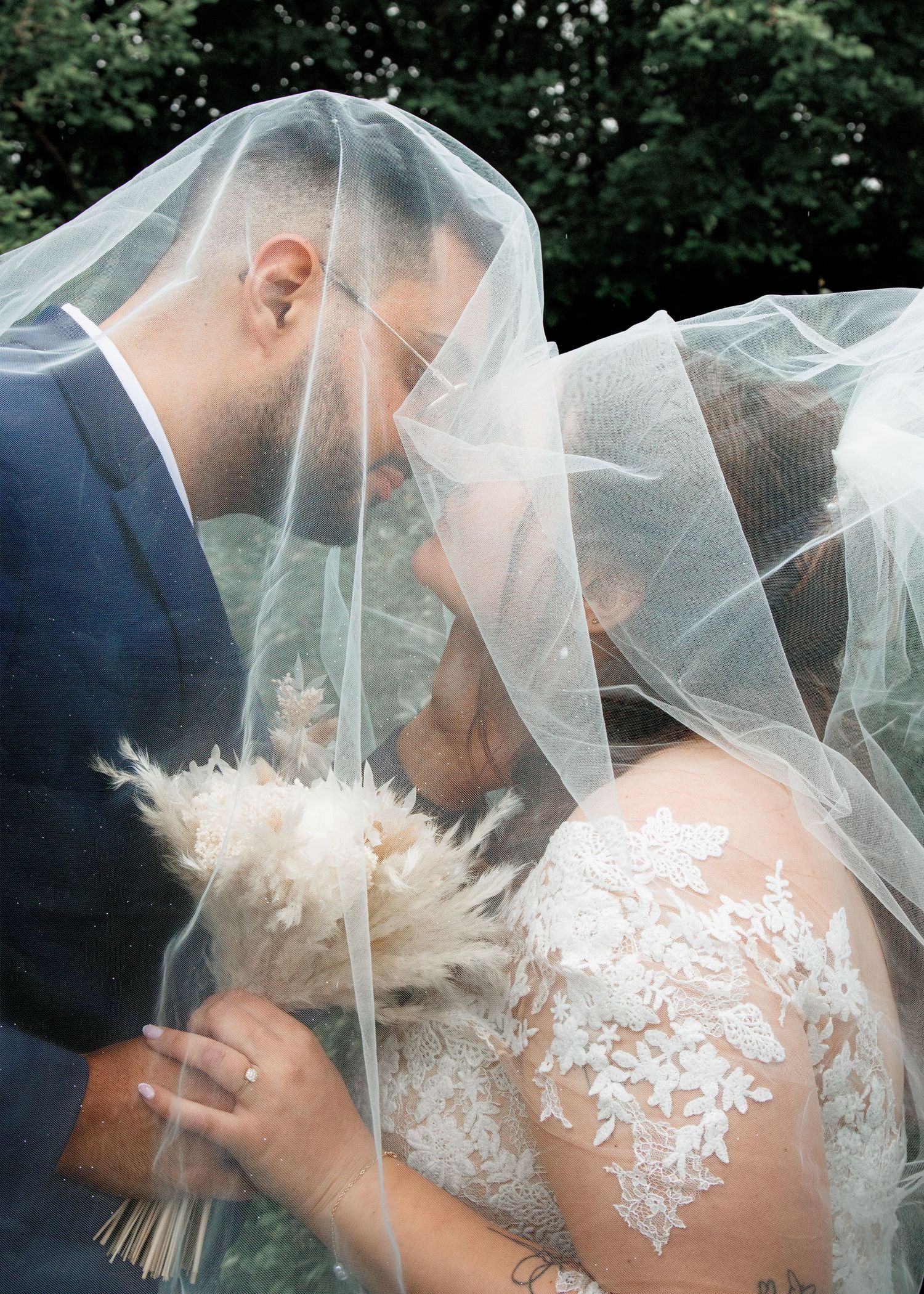 Bride and groom sharing a romantic kiss under a sheer wedding veil, bride holding a boho bouquet in lace gown.