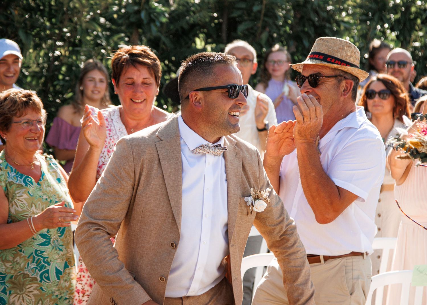 Group of wedding guests laughing together outdoors during a sunny daytime celebration wearing light colored attire.