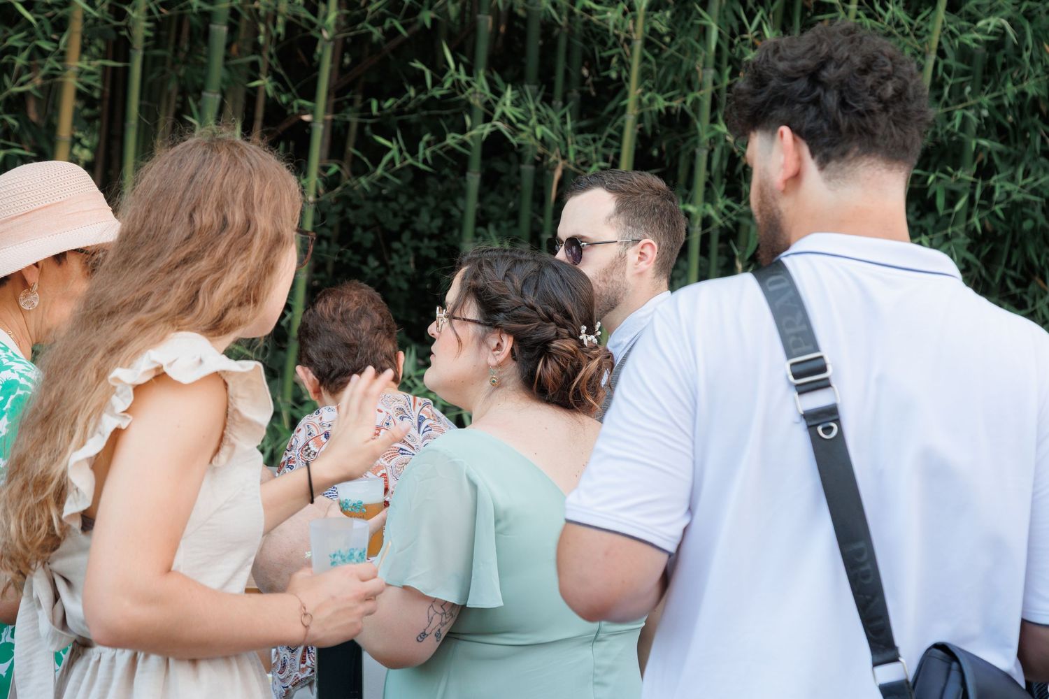 Wedding guests standing in a group outdoors near bamboo, viewed from behind in summer attire.