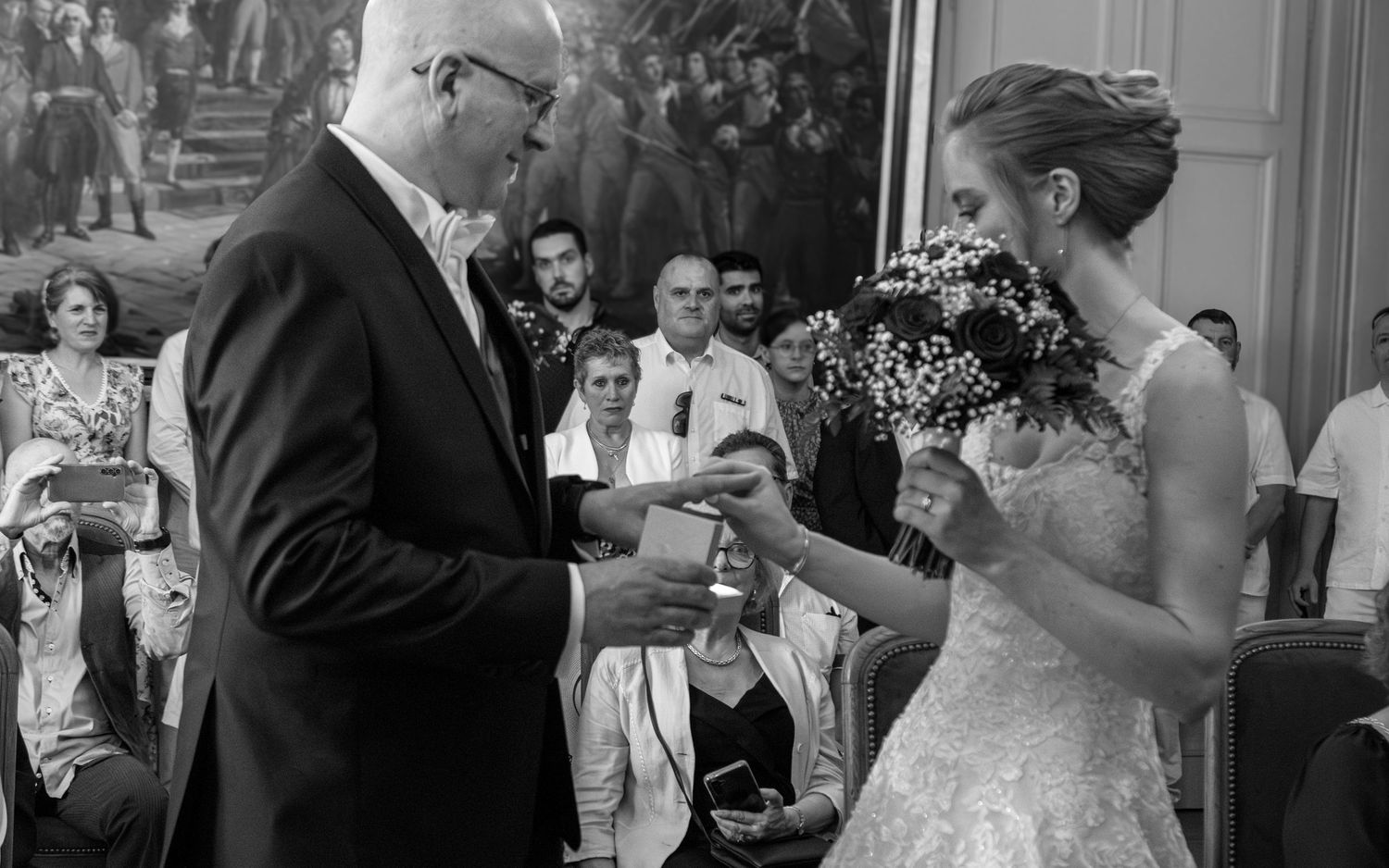Black and white wedding ceremony photo of bride exchanging rings with officiant, guests watching in elegant venue.