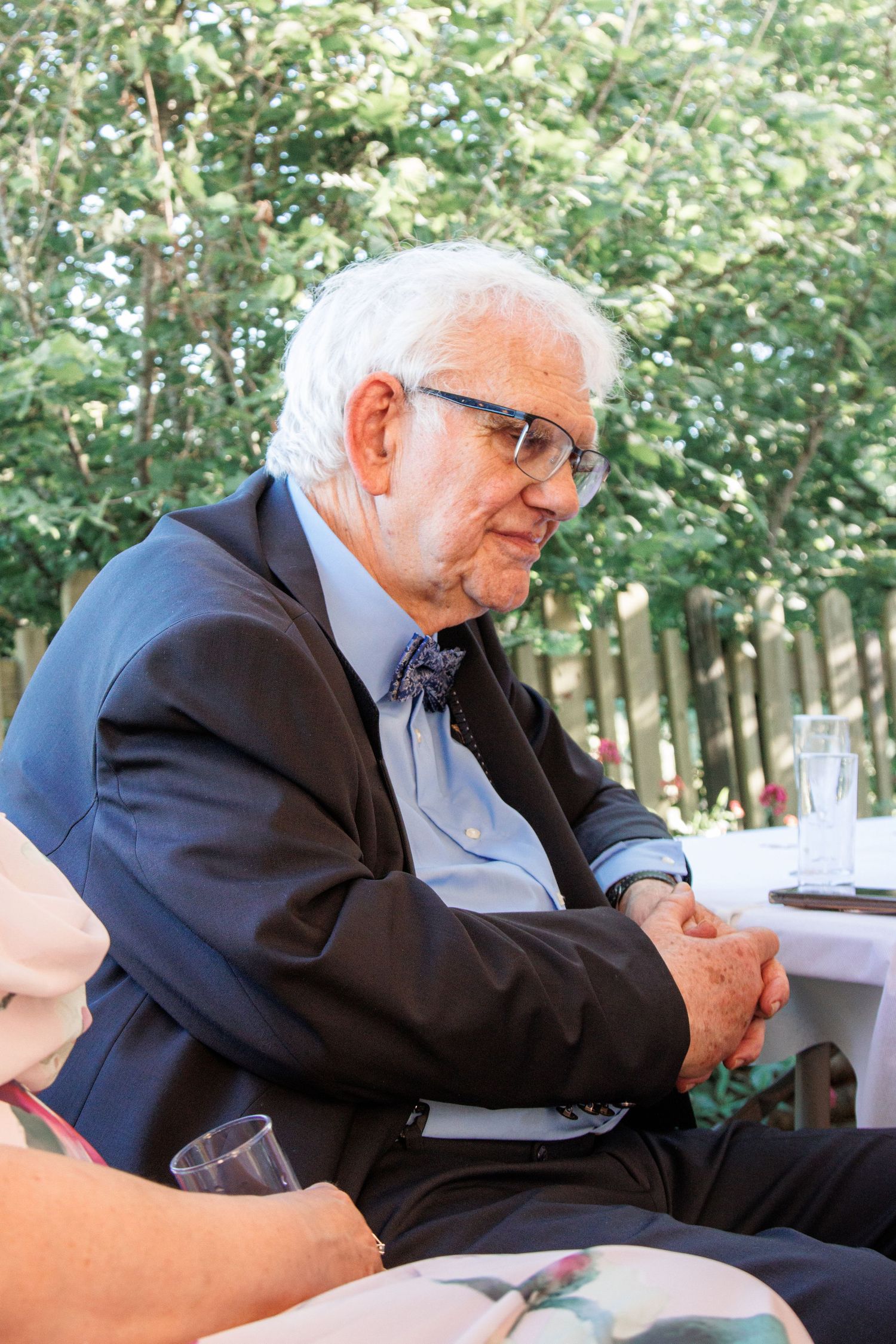 Elderly man with white hair and glasses wearing a dark suit with bow tie seated outdoors at a formal event.