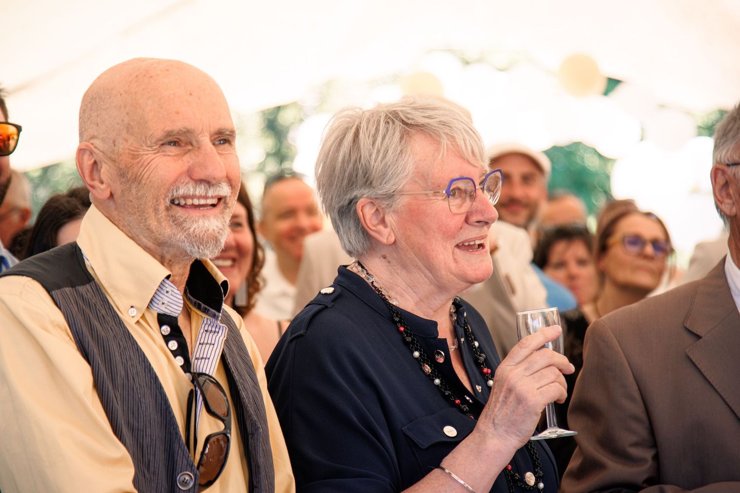 Elderly couple smiling and laughing at a wedding reception under a marquee tent, woman holding champagne glass.
