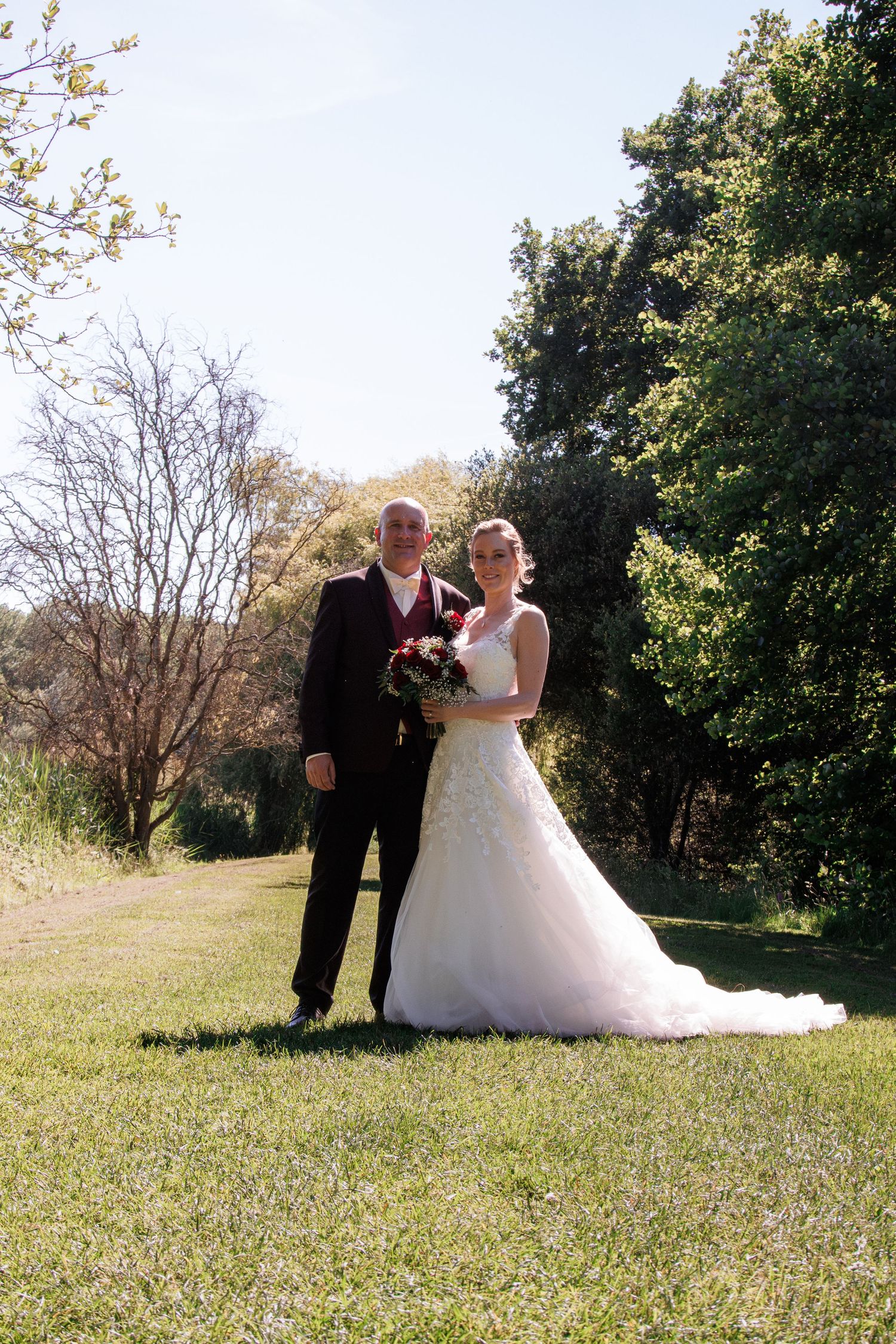 Bride and groom posing together outdoors on green lawn with trees on sunny wedding day.