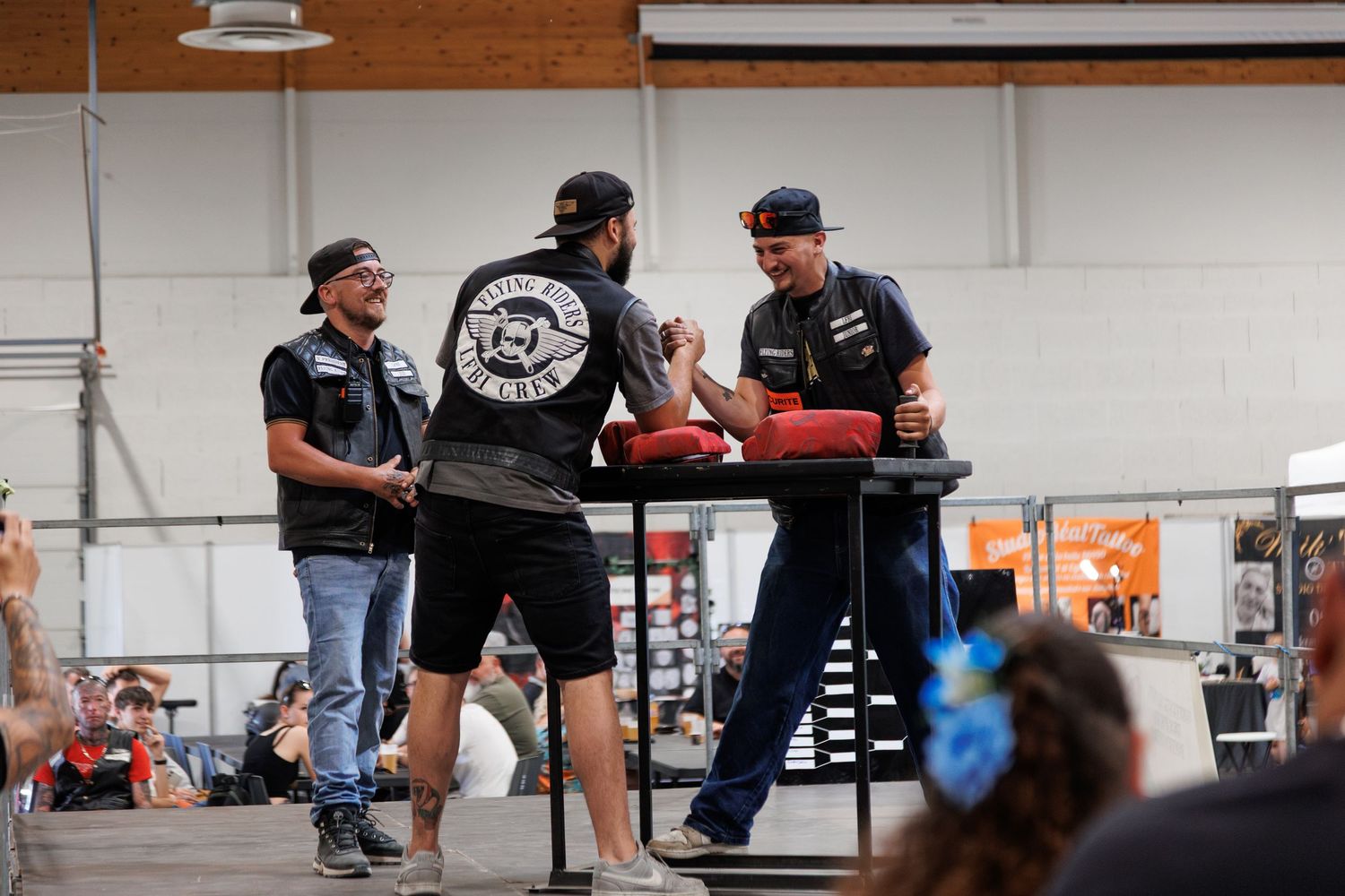 Three men in biker vests arm wrestling on stage at an indoor motorcycle rally event.