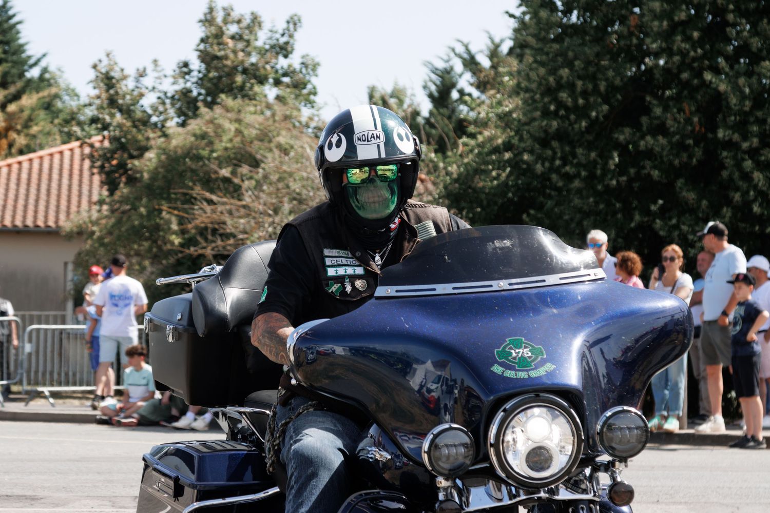 Motorcyclist wearing a Star Wars helmet rides a blue Harley-Davidson at an outdoor event on a sunny day.