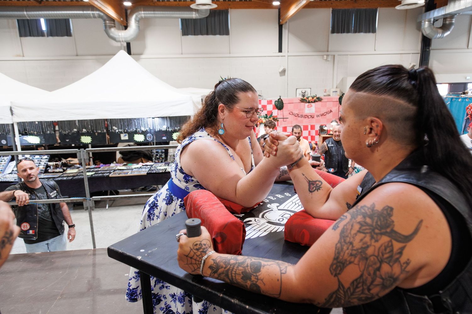 Two tattooed women arm wrestling at an indoor tattoo convention event with vendors in the background.