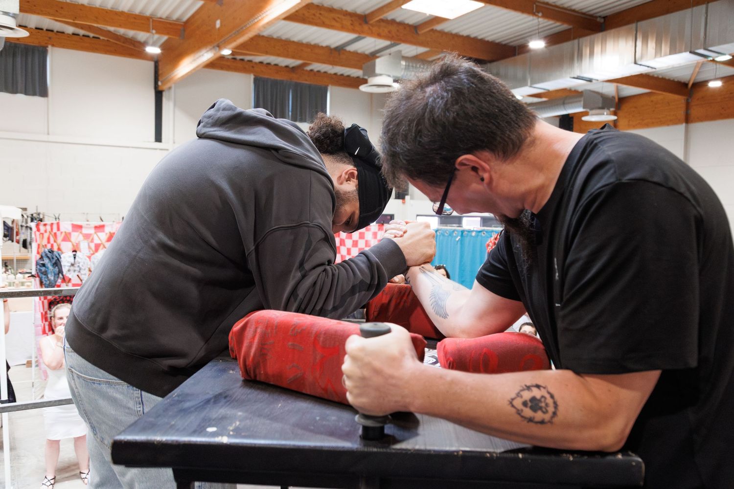Two tattoo artists carefully working on a red sleeve tattoo in a busy convention hall setting.