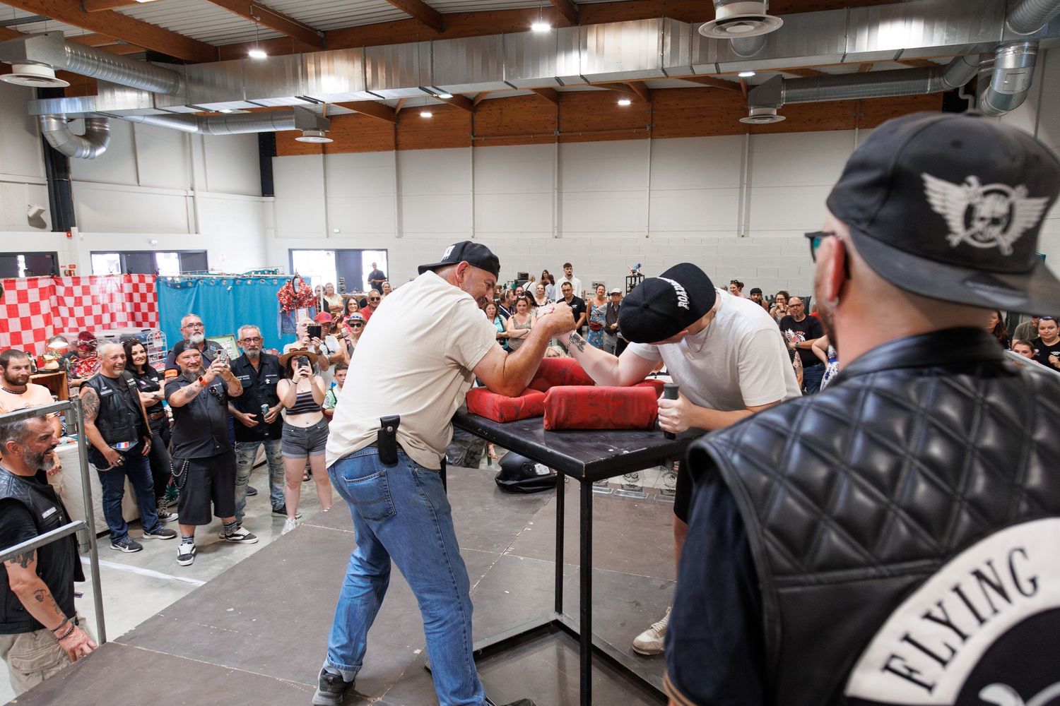 Two men arm wrestling at a competitive event with a crowd watching inside an indoor venue.